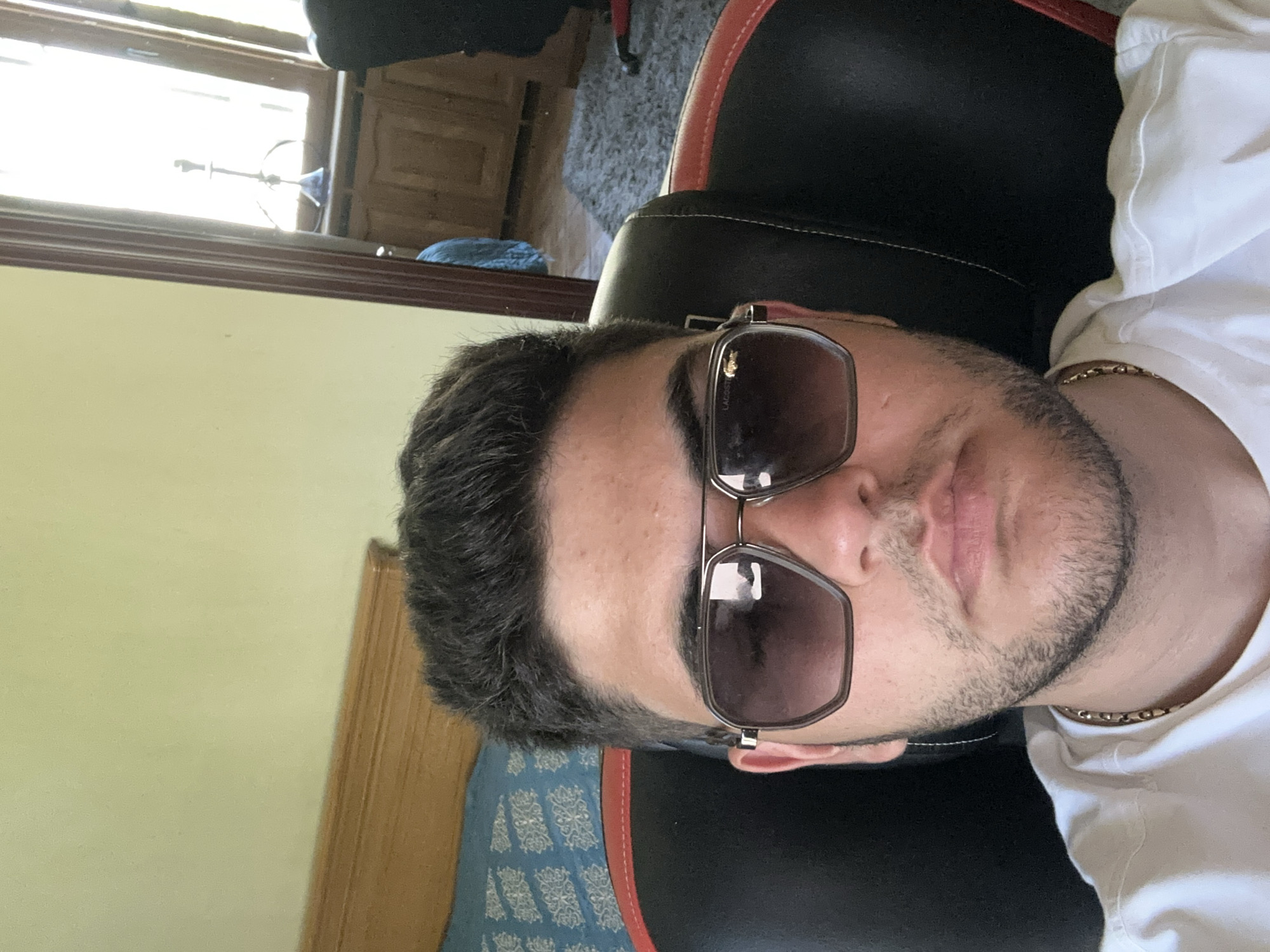 A close-up, vertical shot shows a man wearing sunglasses and a white t-shirt, positioned in a black and red gaming chair. The man has dark hair and a trimmed beard. Part of a gold chain is visible around his neck. The background includes a window with a reflected object, a wooden cabinet, a blue cushion, and a carpeted floor. The setting is most likely indoors. The location is not identifiable as Yerevan, Armenia from this image.