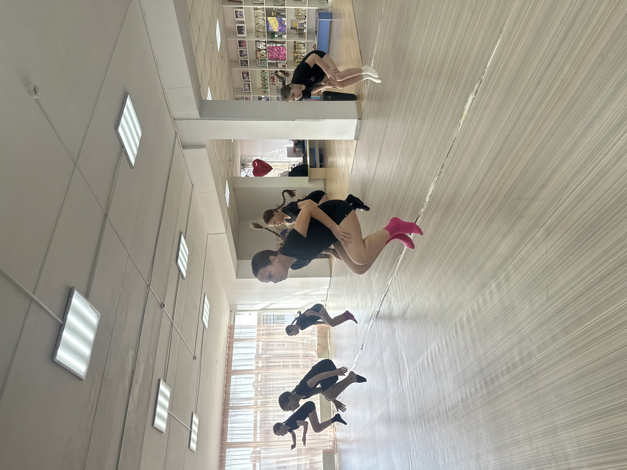 The image shows an indoor dance studio in Votkinsk, Russia. Five girls, likely students, are practicing a dance routine. They are dressed in black outfits and socks of varying colors: white, black, and pink. The girls are arranged in a line, seemingly mirroring each other's movements. The studio has a light-colored wooden floor, white walls, and rectangular fluorescent lighting fixtures on the ceiling. In the background, there is a trophy shelf, a window, and a red heart-shaped balloon.
