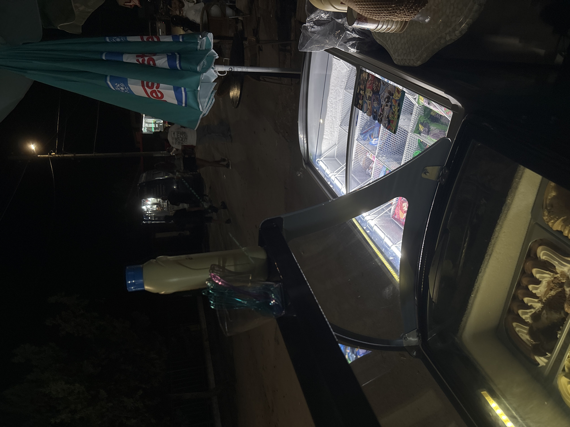 The image depicts an outdoor commercial scene at night in Kostievo, Bulgaria. In the foreground, an illuminated ice cream display freezer with a partially open curved glass lid reveals packaged ice cream products and a branded "NUII" menu. To the left, a second, lower freezer section contains scoopable ice cream. On a dark counter surface to the left, a yellow plastic bottle with a blue cap stands upright next to a clear container holding multiple colorful, twisted plastic utensils. To the immediate right of the display freezer, a beige, textured object, possibly a woven hat, rests on a white, textured surface, adjacent to a crumpled clear plastic bag.
In the midground, several individuals are visible walking on a paved street; one person is seen from behind wearing a white "The North Face" t-shirt. A large, teal market umbrella, folded and displaying a white "es" logo with a snowflake graphic, occupies the right side of the midground. The background is dark, revealing dark trees to the left, distant structures, and multiple artificial light sources, including a prominent street lamp emitting a bright glow. The overall scene indicates a late-evening vendor operation in a populated area.