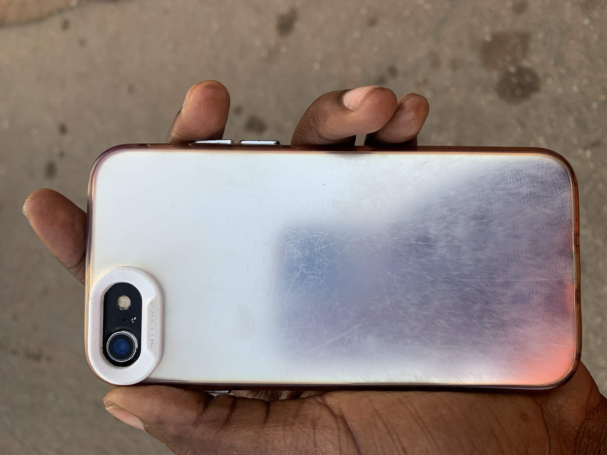 A person's hand is holding a smartphone, likely an iPhone, with the back facing the camera. The phone has a protective case with a white border around the camera area and a rose-gold or copper colored edge. The back of the phone is scratched and shows some wear. The person's hand is dark-skinned and the fingers are visible holding the phone. The background is a blurry gray surface, possibly concrete or asphalt. There are no visible indications suggesting that the picture was taken in New Stadium, Nigeria.