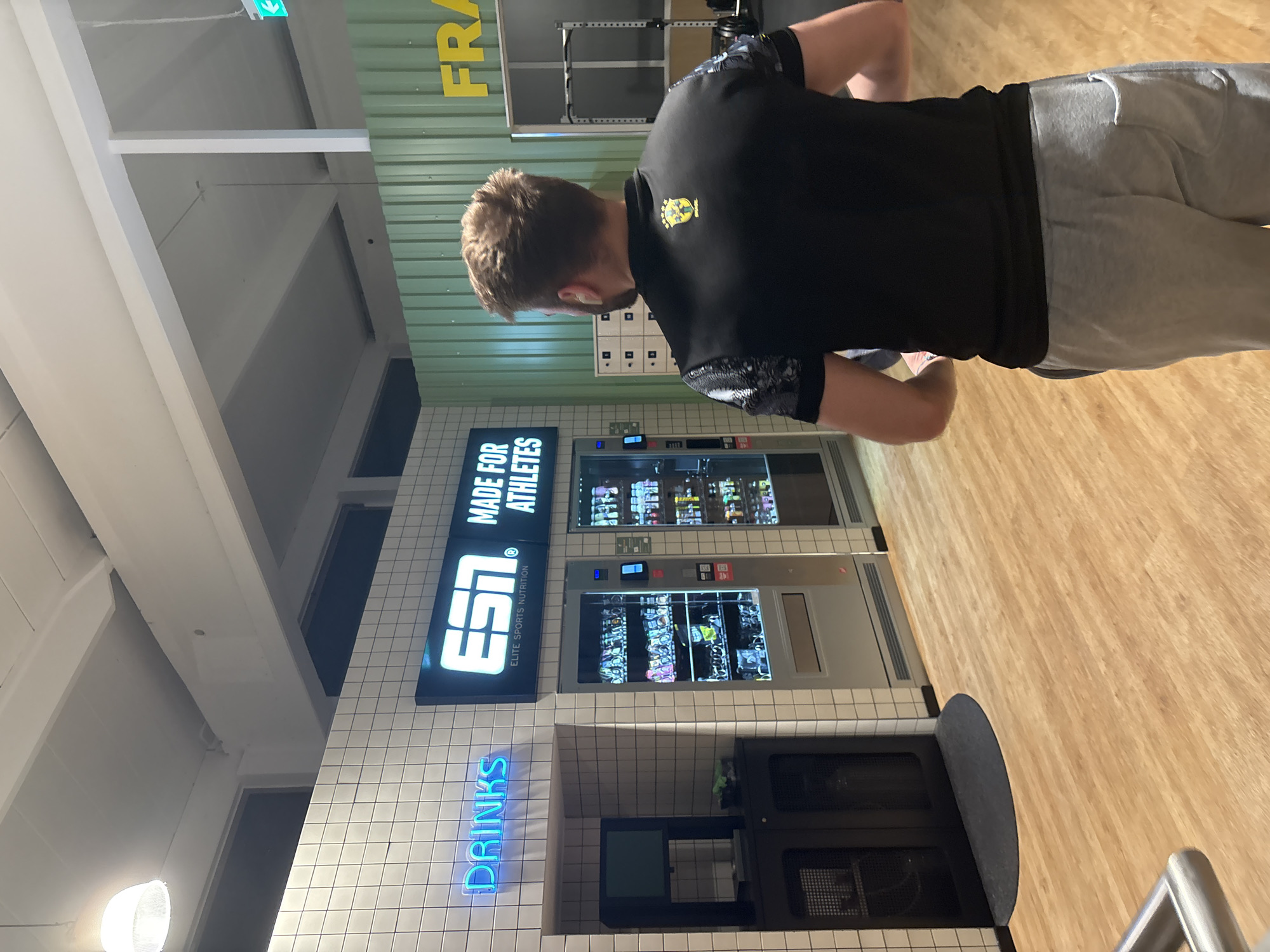 A man stands in front of vending machines in Mönchengladbach, Germany. He is wearing a black shirt and gray sweatpants. The vending machines sell drinks and other products. The sign above the machines reads "Elite Sports Nutrition."