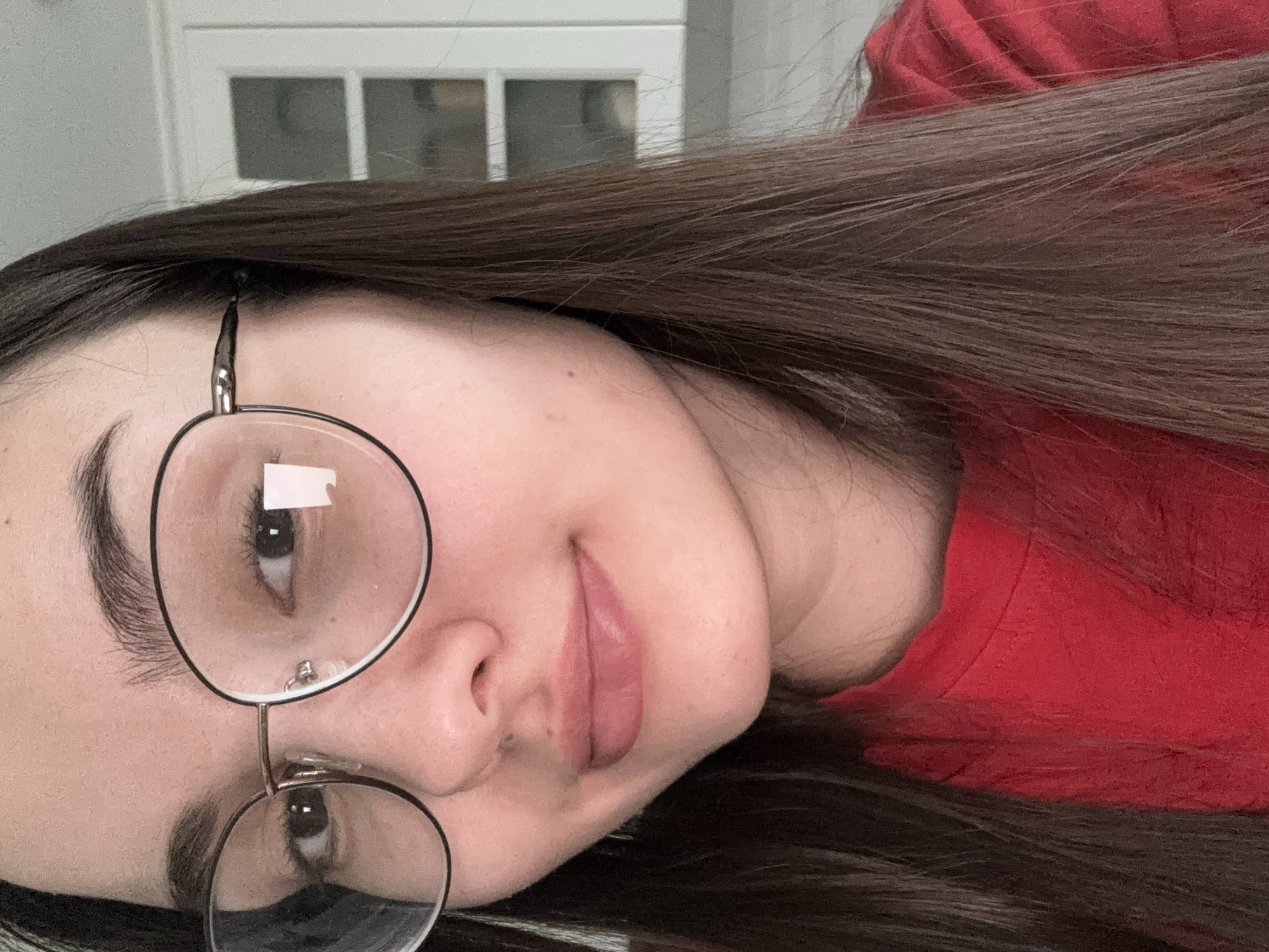 A close-up, angled headshot of a woman with long, straight dark brown hair and light skin. She is wearing round, thin-rimmed glasses. Her expression is neutral with slightly pursed lips. She is wearing a red shirt. The background is blurred, showing a white wall and a window with a grid pattern. The location appears to be indoors. There is no evidence in the image to suggest this image was taken in Chaikovsky, Russia.