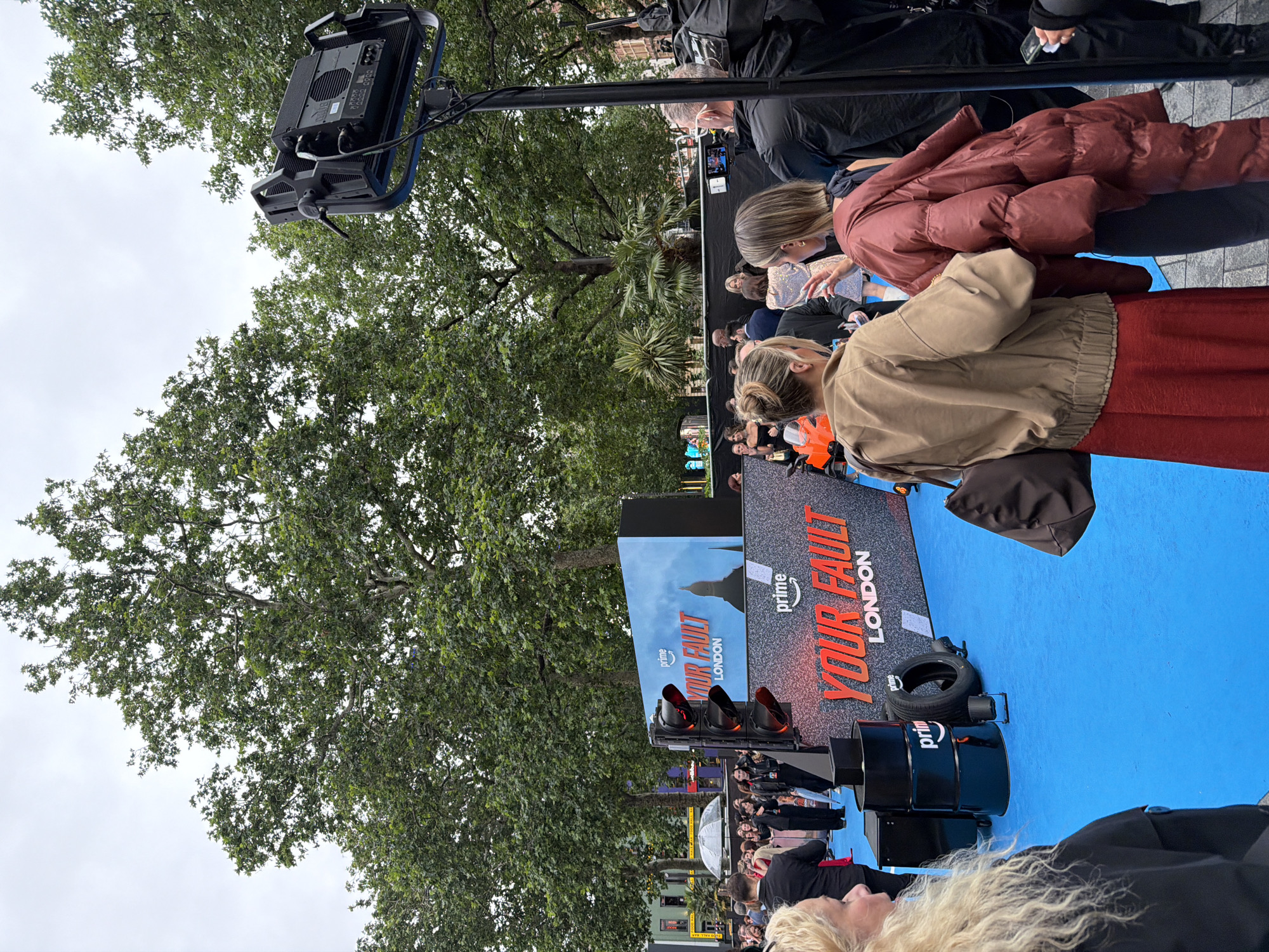This is a medium-shot, eye-level, full-color photograph taken outdoors at a red carpet event for "Your Fault London," likely at a film premiere or promotional event. The scene is set on a bright blue carpet under a cloudy sky, with lush green trees forming a backdrop.
In the foreground, a person with long blonde hair is visible on the left, facing away from the camera. To their right, two individuals are talking, with one wearing a tan jacket and a red skirt, and the other a puffy red coat. In the middle ground, a large black traffic light fixture is positioned to the left of a screen displaying the "Your Fault London" logo and imagery. Several people are gathered around, some appearing to be photographers or media personnel, and others attending the event. Black barrels and a tire are also present on the blue carpet.
In the background, more trees and buildings are visible, suggesting an urban setting within London. The overall atmosphere suggests a lively event with a focus on media coverage and public engagement.