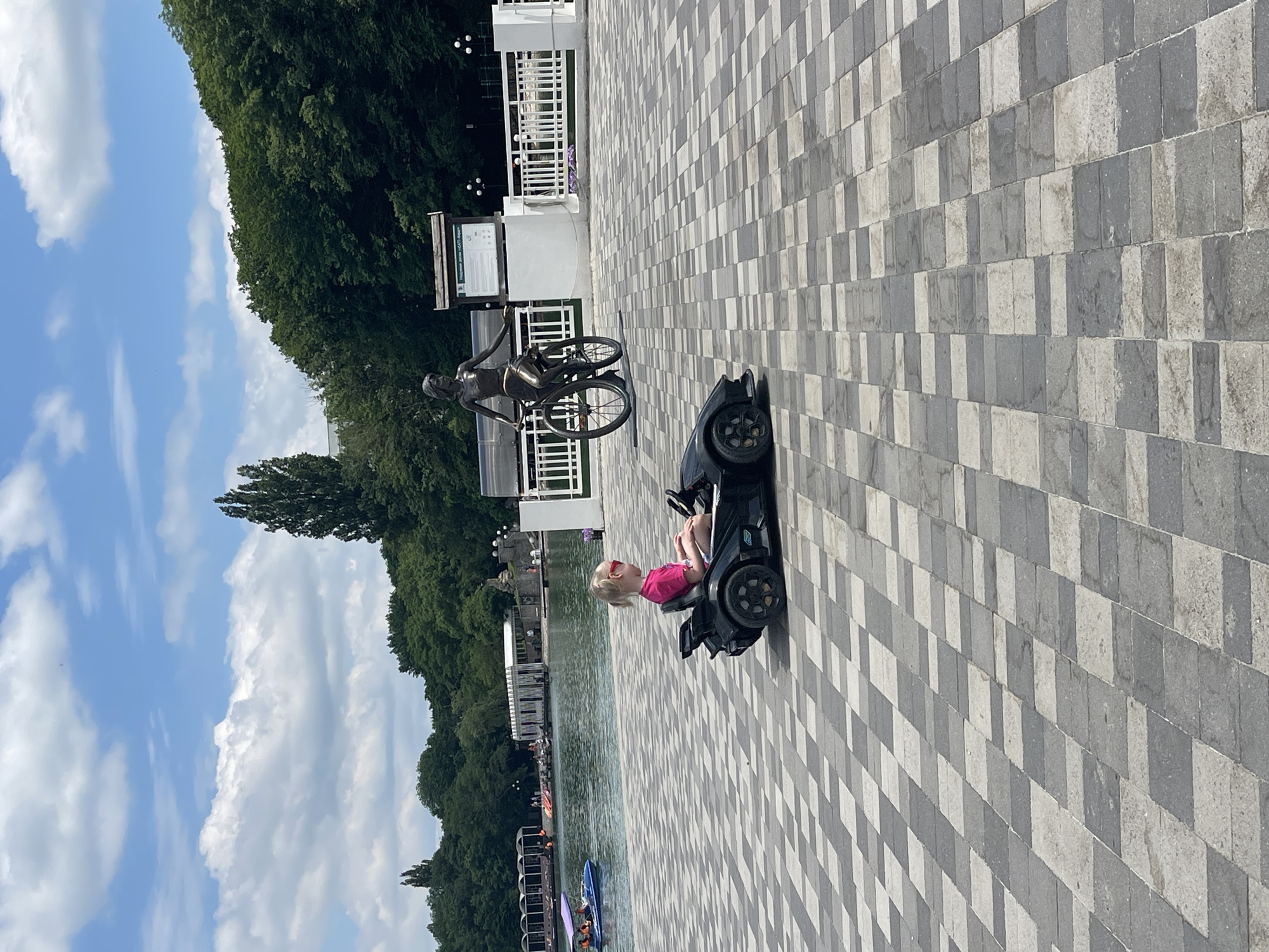 In the city of Zheleznovodsk, Russia, a young girl with blonde hair and pink sunglasses is seated in a black toy car on a checkered paved area. She is wearing a pink t-shirt and white shorts. Behind her, a bronze statue of a woman riding a bicycle stands on a platform. In the background, a body of water is visible, with several boats and people enjoying recreational activities. Lush green trees and a cloudy blue sky complete the scene.
