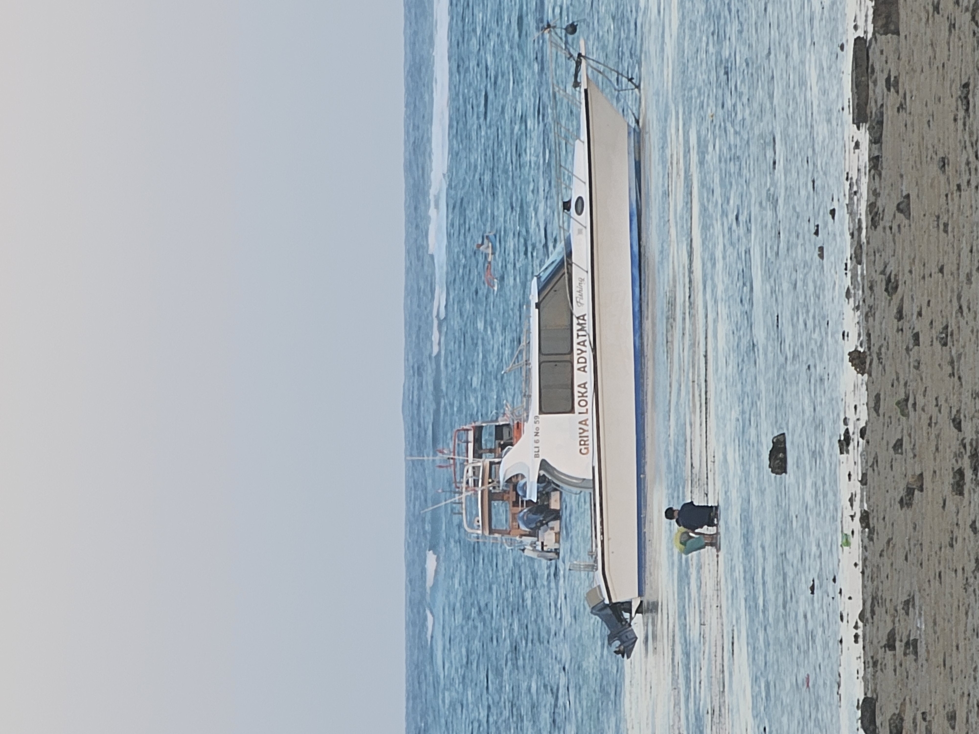 In the shallow waters off the coast of Denpasar, Indonesia, a white motorboat with blue trim is anchored near the shore. The boat, identified by the text "BLI 6 No 59 GRIYA LOKA ADYATMA Fishing" on its side, has an outboard motor at its stern. In the foreground, two figures, likely children, are wading in the water. One child, wearing a yellow shirt, is partially submerged, while the other, dressed in dark clothing, is kneeling. The water is calm, with small waves visible in the distance, and the seabed is covered with rocks and sand. Another boat with a higher cabin structure is moored behind the main vessel, and a lone figure on a surfboard can be seen further out at sea. The sky is overcast with a pale, hazy light.
