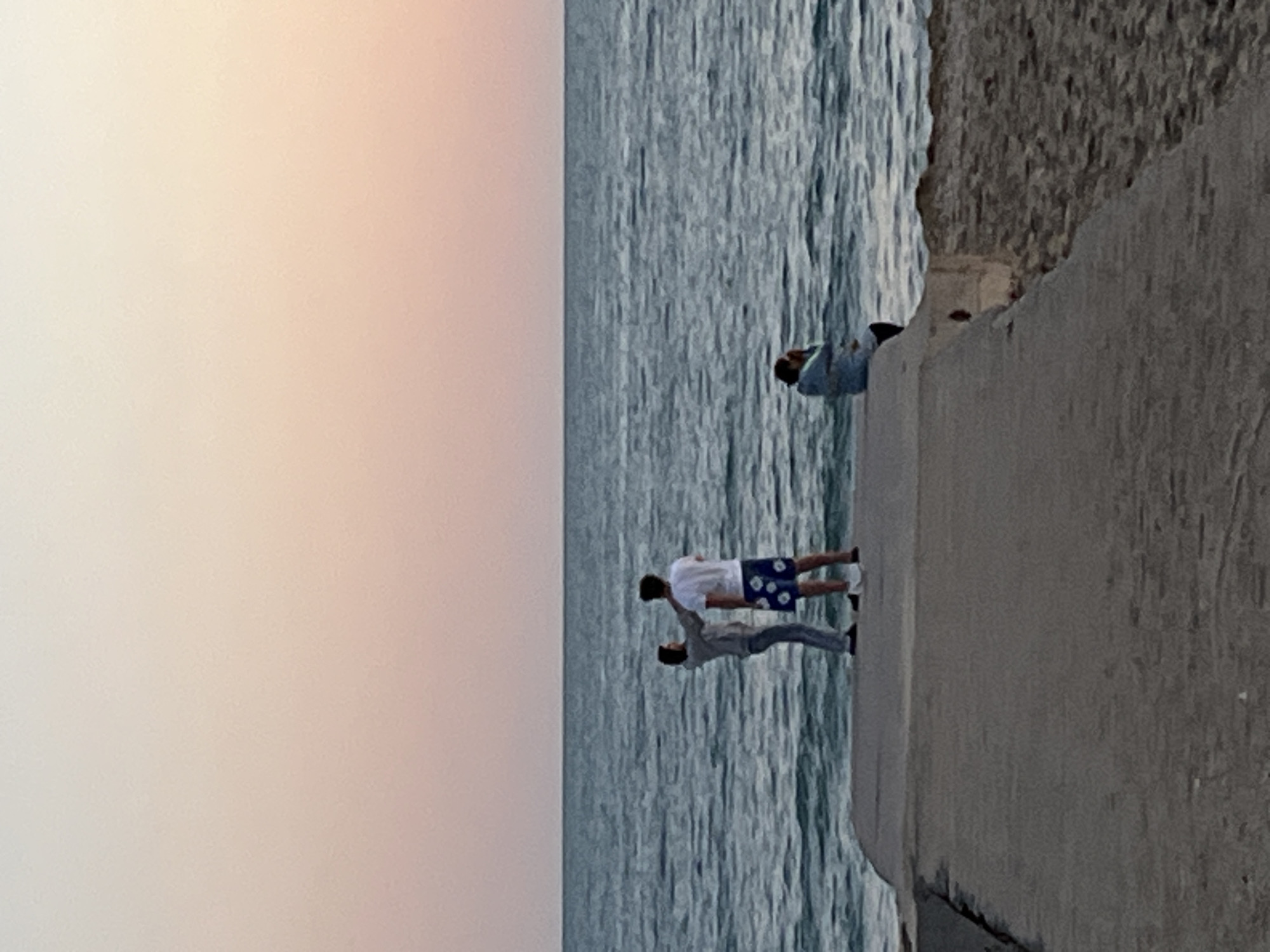 The image depicts a seaside view in Alanya, Turkey, with a concrete wall or structure forming the edge of the scene on the right. Two individuals stand near the edge of the wall, with one person in a white shirt and blue patterned shorts, and another in a gray shirt and blue jeans. They appear to be looking towards the sea. The sea, with visible wave patterns, occupies the central part of the image. In the upper part of the image, a pink sky occupies the space above the water. Another person is seen sitting on the concrete structure further down.