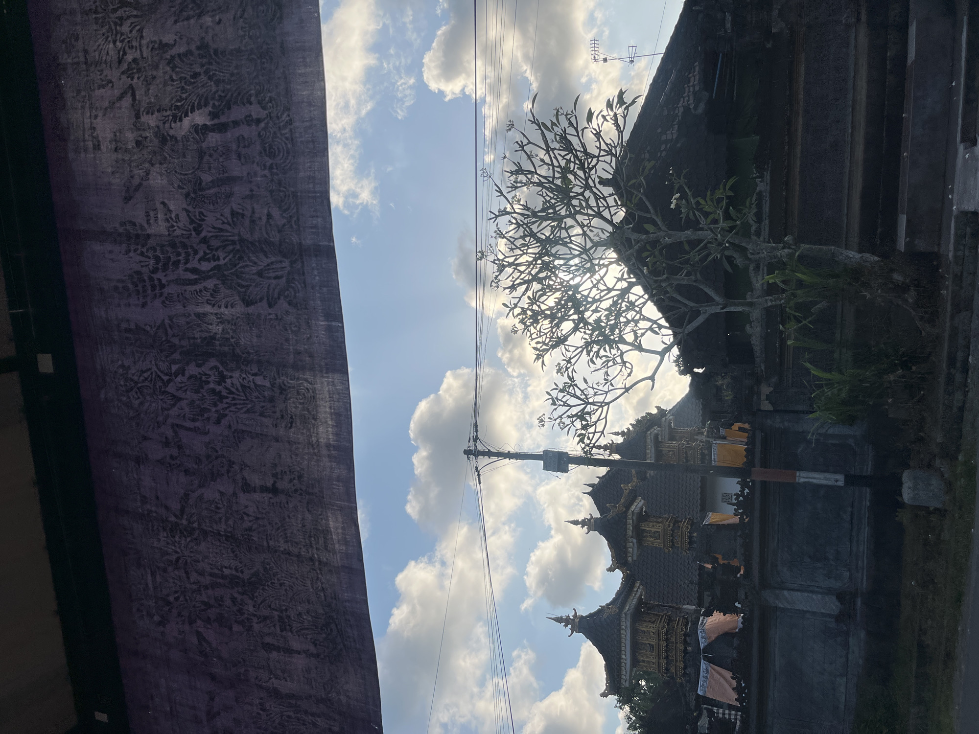 An outdoor, eye-level shot shows a view of a temple in Mendoyo, Indonesia, partially obscured by a dark purple, patterned fabric hanging in the foreground. The sky is blue with scattered white clouds and the sun is shining. In the mid-ground, a traditional Balinese temple structure with dark, tiered roofs and ornate gold embellishments is visible. A stone wall with decorative carvings surrounds the temple complex. A bare-branched tree with a few green leaves stands in front of the temple. Power lines and a utility pole are also present. The lower part of the image shows a dark, grassy area and a street.