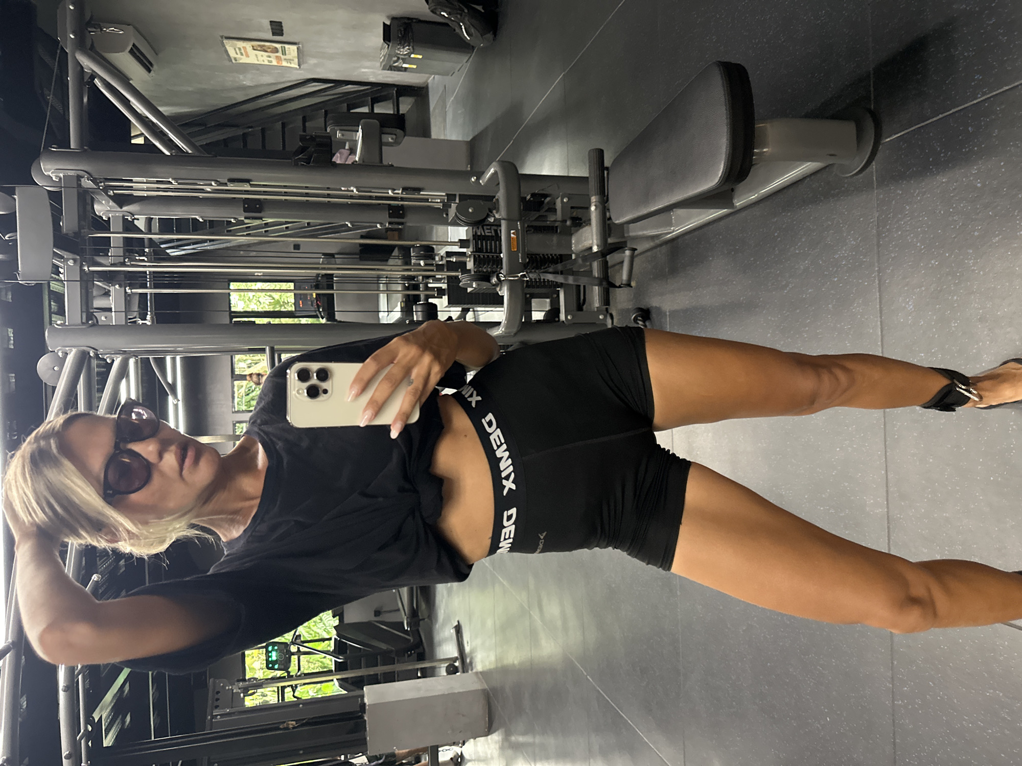 The image shows a woman taking a selfie in a gym. She is wearing a black t-shirt, black shorts with a white logo that says "DEWIX", and sunglasses. Her hair is blonde and she has her left hand on her head. In the background, there is gym equipment, including a weight machine and a bench. The floor is gray and there is some greenery visible through a window. The lighting suggests it is daytime. The woman's expression appears neutral. The text "DEWIX" is visible on her shorts. The location is identified as Mengwi, Indonesia.