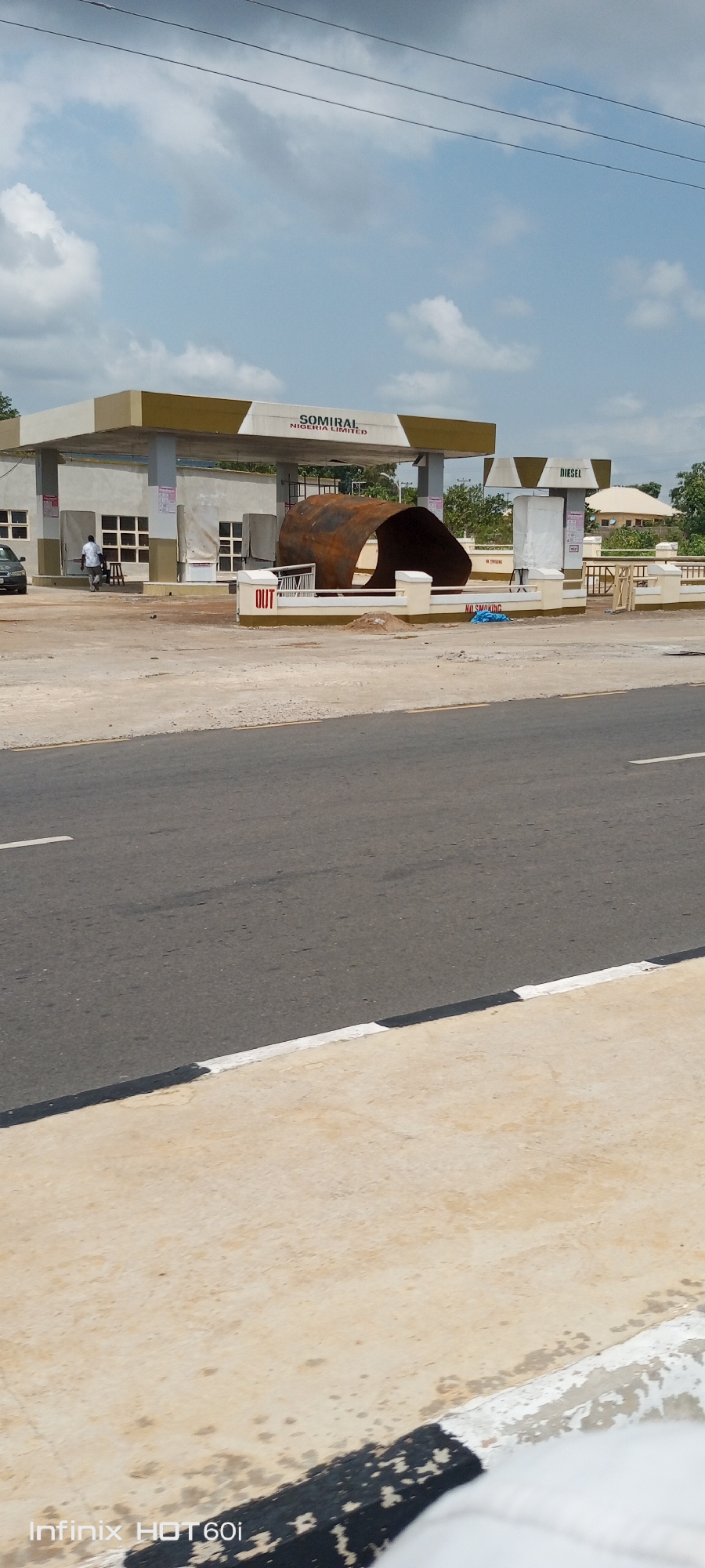 The image depicts a fuel station, SOMIRAL NIGERIA LIMITED, in Zango-ndaji, Nigeria, during the day. The station has a standard design with fuel dispensers under a canopy, and a sign indicating "DIESEL". A large, rusted cylindrical object, possibly a section of a tank or pipe, rests on the ground in front of the station. The ground around the fuel dispensers appears to be under construction or recently renovated. A man in light-colored clothing stands near a car next to the station. The foreground features a road with a white line marking the edge. The sky is partly cloudy.
