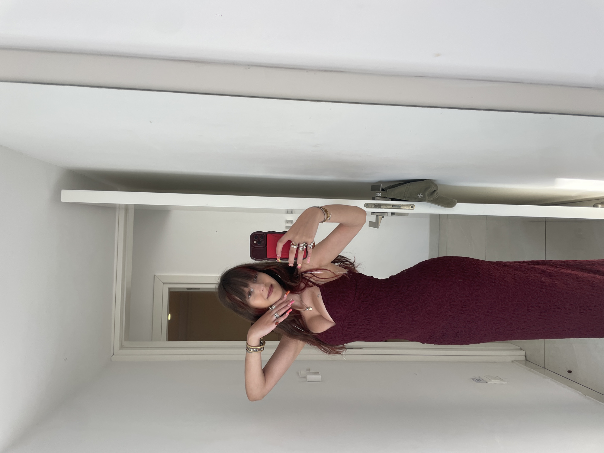A young woman with long, dark brown hair and bangs stands in a hallway, taking a mirror selfie with her phone. She is wearing a long, form-fitting, dark red lace dress with a V-neck and thin straps. She is accessorized with several rings on her fingers, bracelets on both wrists, and a necklace with a heart-shaped pendant. Her nails are painted pink and decorated. The hallway appears to be in a modern residential building, with white walls and tiled flooring. A doorway is visible in the background, leading to another room. A dark green cap is hanging on a hook on the doorframe to the right. The lighting suggests it is daytime. The scene does not depict anything related to Paphos Municipality, Cyprus.