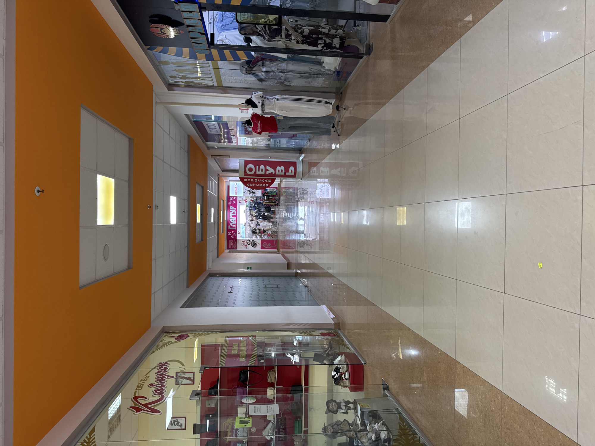 The image depicts the interior of a shopping mall corridor in Yeysk, Russia. The corridor is wide and well-lit, with a high ceiling painted orange. The floor is tiled with large, light-colored ceramic tiles, reflecting the overhead lights. On either side of the corridor are storefronts with glass facades, displaying various goods.
On the left, a shoe store is visible with mannequins displaying footwear and handbags. The store's signage is in Russian, featuring a decorative script. Further down, other shops exhibit clothing and accessories.
On the right, several storefronts are present. One shop displays mannequins dressed in casual athletic wear, with branding that appears to be Adidas. Another shop showcases fashion items behind glass. In the distance, more stores and shoppers can be seen. The overall atmosphere suggests a typical commercial environment.