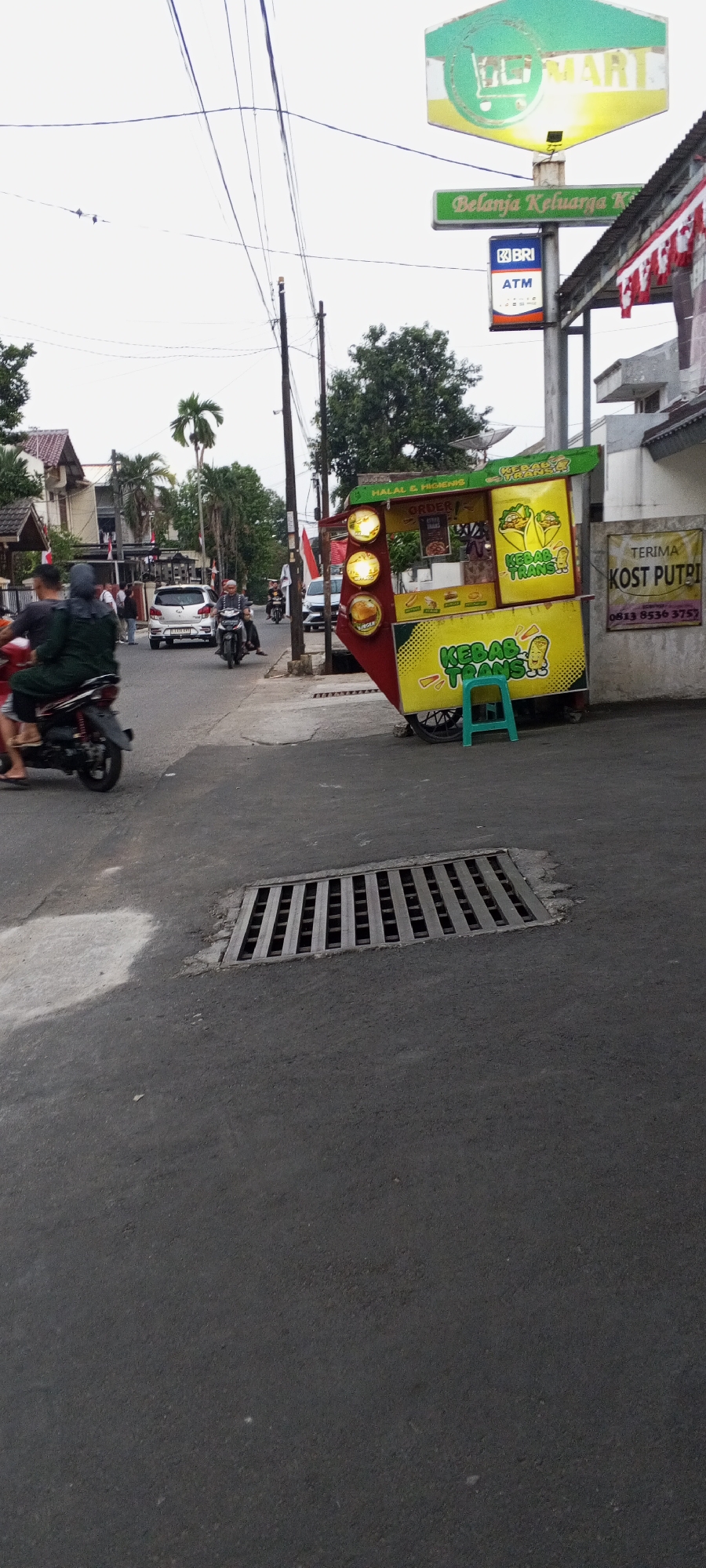 The image depicts a street scene in Depok, Indonesia, with a focus on a Kebab Trans street food cart. The cart is brightly colored in yellow and red, with images of kebabs and a cartoon character. Above the cart, a sign reads "HALAL & HIGIENIS" and "ORDER". To the right of the cart, a yellow and white sign advertises "TERIMA KOST PUTRI" with a phone number. A small green stool is placed in front of the cart.
In the background, traffic moves along the asphalt road. Several motorcycles are visible, including one carrying two people in the foreground. A white car is also present on the road. The environment includes trees, buildings, and utility poles with numerous overhead wires. A large metal grate covers a drainage system in the road in the foreground. A partial view of a BRI ATM sign is visible on a pole above the street food cart. The sky is overcast.