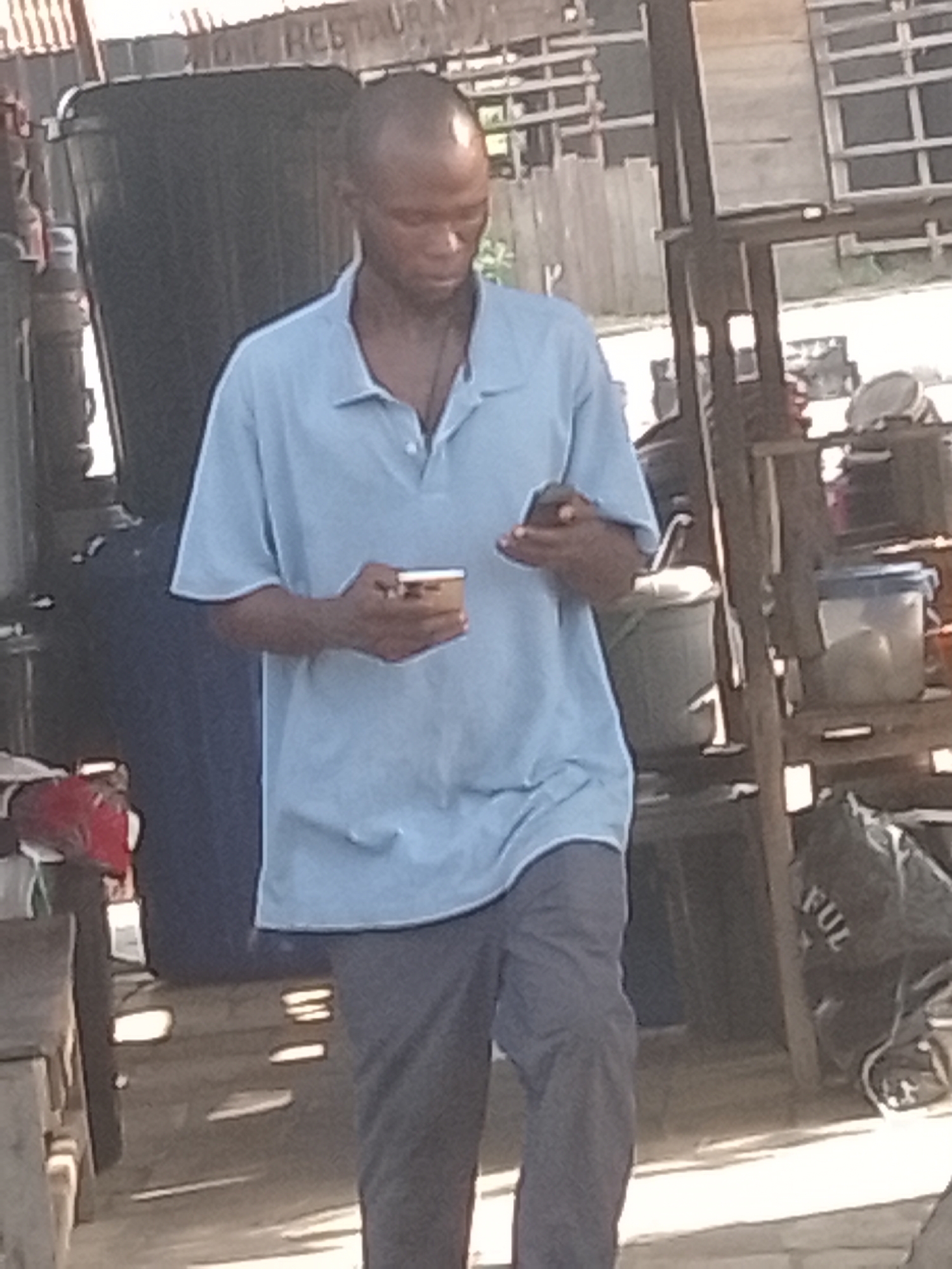 A bald man, appearing to be of African descent, is depicted in what seems to be an outdoor or semi-outdoor commercial area during the day. He is wearing a light blue polo shirt and dark trousers, and his head is bowed as he focuses on two mobile phones he holds. One phone, in his left hand, is a smaller, light-colored device, while the other, in his right hand, is a darker smartphone.
Behind the man, a large, dark plastic barrel or container is visible. To his right, there are various items on rustic wooden shelving, including a light blue and a grey plastic bucket or container. A dark bag with the partial word "FUL" is on the ground to the lower right. To the man's left, a red object, possibly a piece of fabric, rests on a wooden surface. The background features a corrugated metal roof and wooden planks, indicative of an informal structure. Visible on the corrugated roof is the partial text "ONE RESTAURA," suggesting a restaurant or eatery. The bright, even lighting indicates it is daytime.