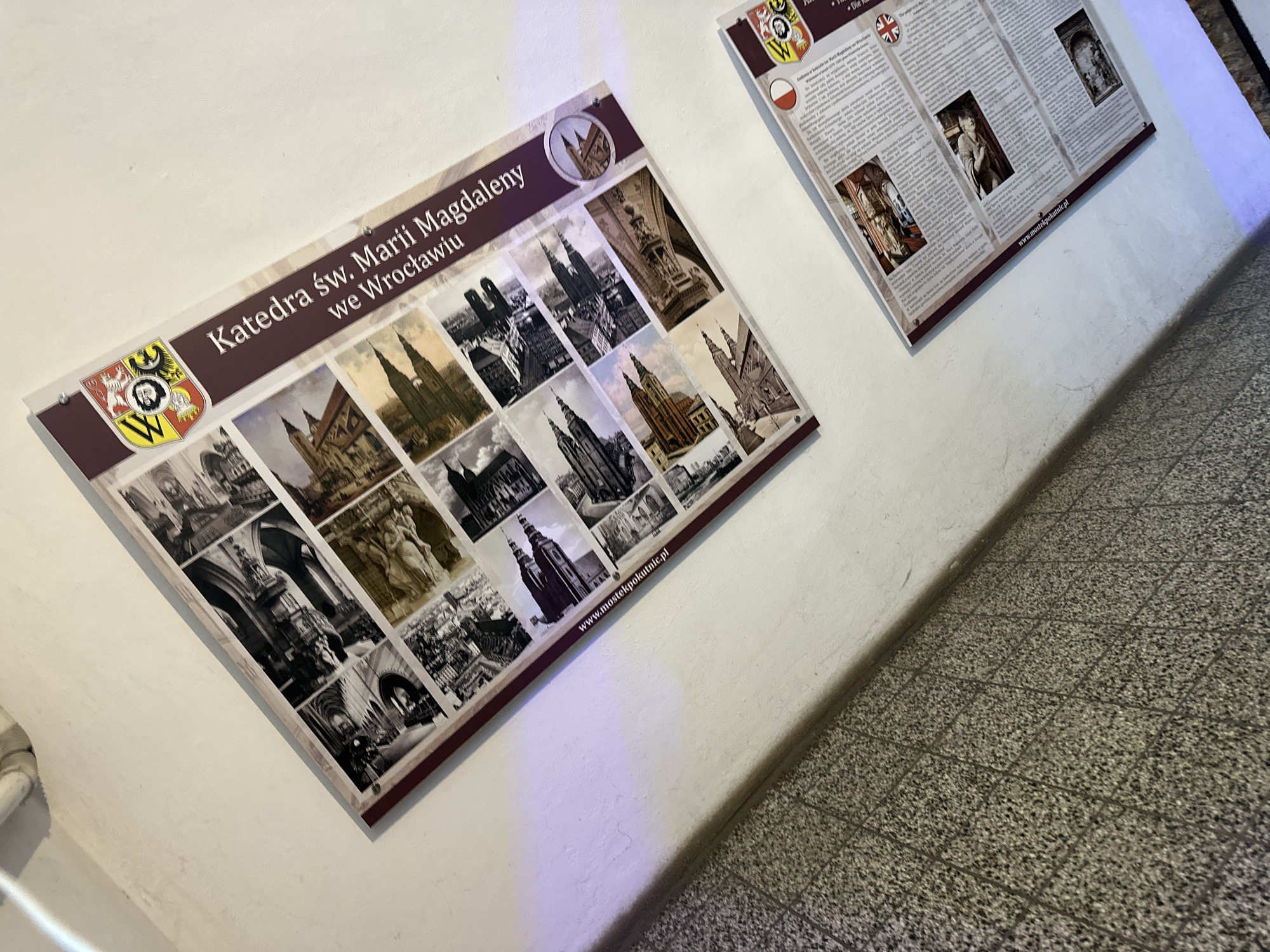 Two posters are mounted on a white wall next to a tiled floor. The poster on the left displays the title "Katedra św. Marii Magdaleny we Wrocławiu" (Cathedral of St. Mary Magdalene in Wrocław) above a collage of grayscale images of the cathedral. It also features a crest with a stylized "W" and the website address "www.mostekpokutnic.pl". The poster on the right is a white information board with text in Polish, English, and possibly other languages, accompanied by images. The scene appears to be inside a building, likely near the cathedral.