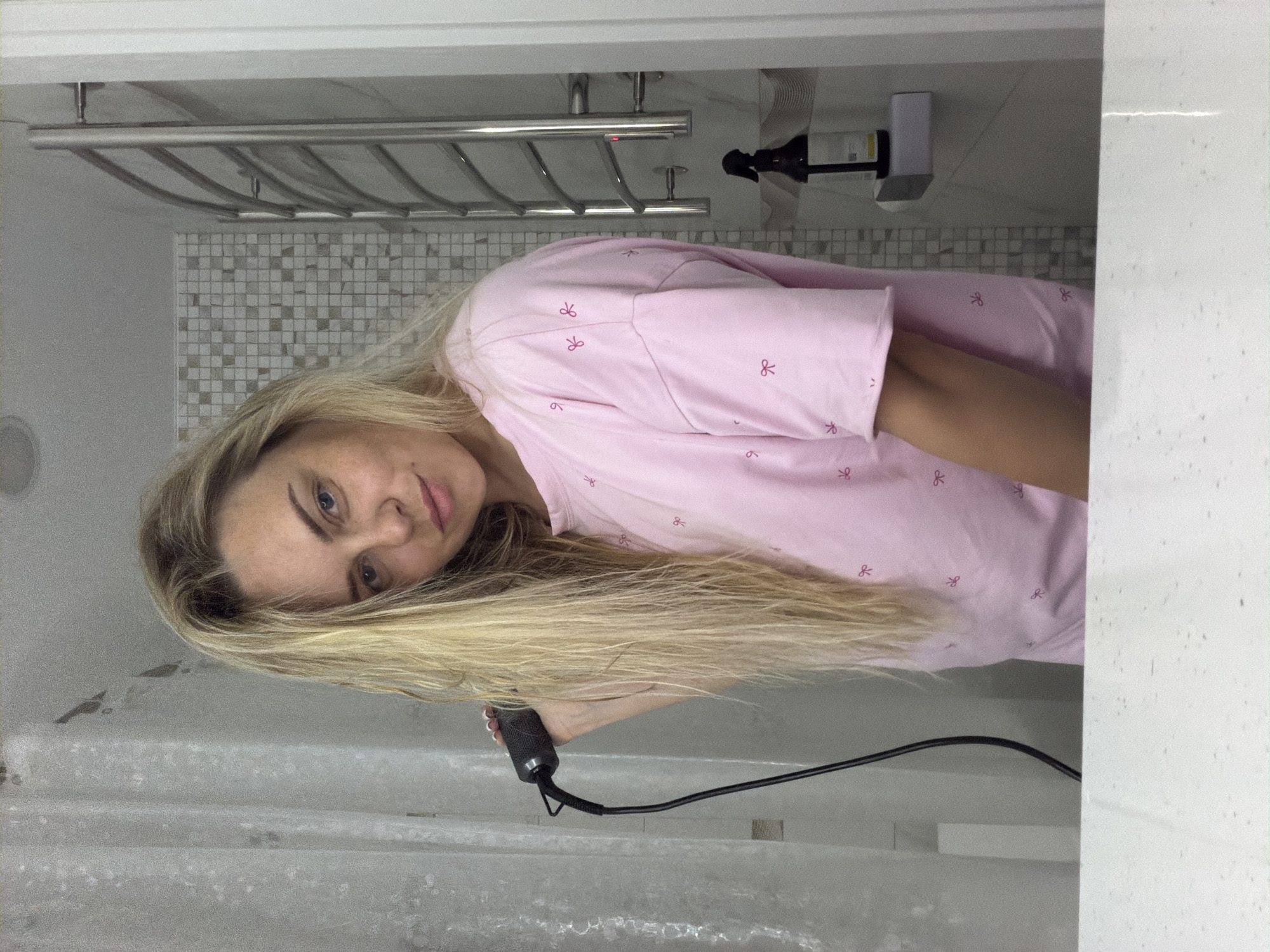 This media file captures a woman taking a mirror selfie in a contemporary bathroom, located in Votkinsk, Russia. The woman has long, straight blonde hair and striking blue eyes, looking directly at the camera with a neutral to slightly pleasant expression. She is dressed in a light pink top or pajama shirt, featuring a repeating pattern of small, dark pink bow-tie symbols.
In her right hand, she holds a black hairdryer with a black power cord trailing downwards. The background reveals a clean and modern bathroom interior. A silver-toned, multi-bar towel rack is mounted on the wall. Below and around it, the wall is decorated with small, square mosaic tiles in a gradient of white, grey, and beige tones. To the right of the towel rack, a wall-mounted dispenser, likely for liquid soap or shampoo, is visible in black and white.
In the foreground, the edge of what appears to be a white bathtub or shower unit is seen on the left, while a white countertop or ledge occupies the right side of the frame. The scene suggests a personal moment, possibly during a morning or evening grooming routine such as hair drying. No discernible text is visible on any objects within the image.