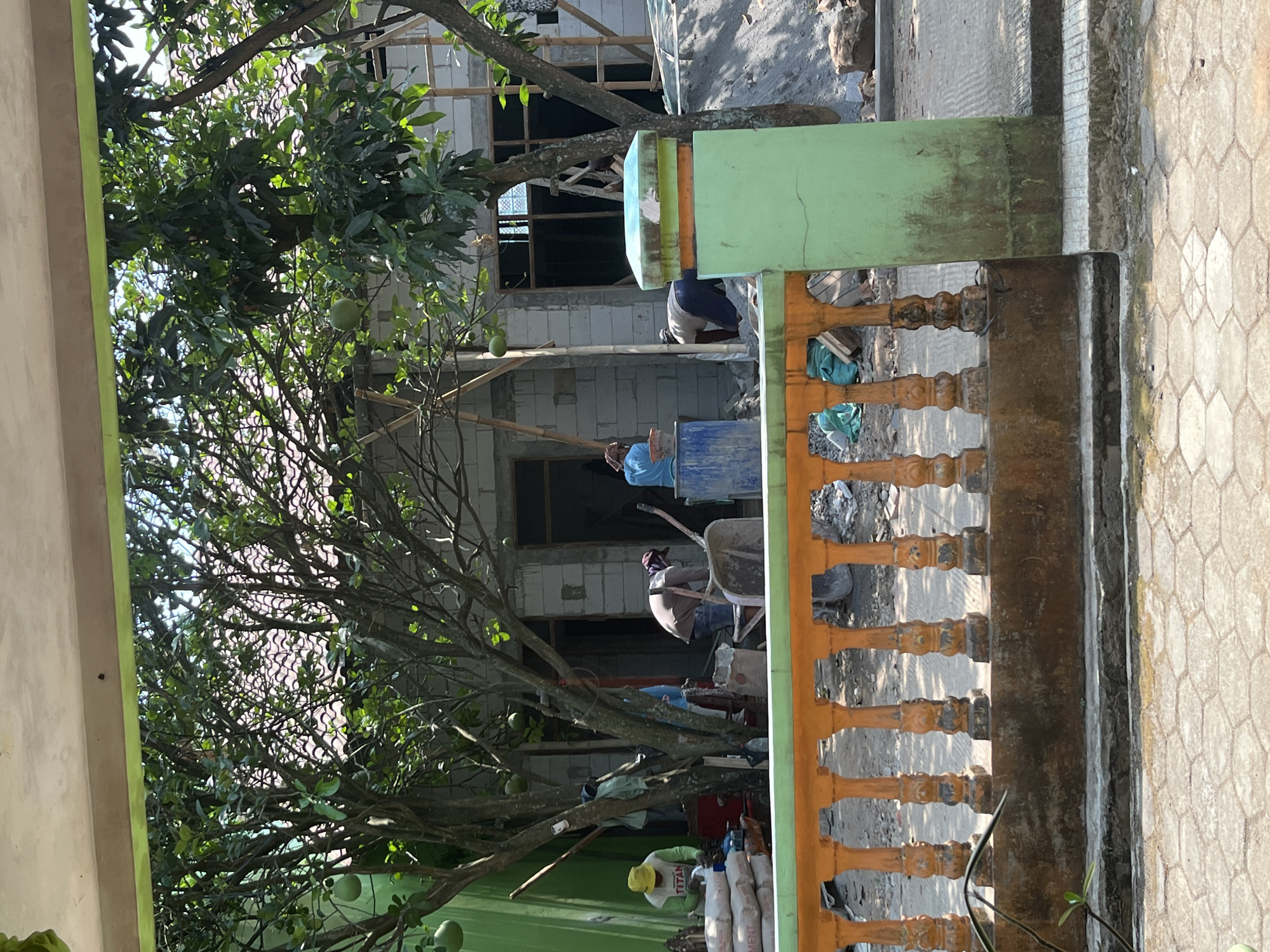 The image depicts a construction scene in Glagah Kulon, Indonesia, under a bright, sunny sky. In the foreground, a weathered, orange-colored railing with decorative balusters separates the viewer from the construction site. A portion of a green pillar stands to the right of the railing, showing signs of age and moss growth. Behind the railing, several workers are engaged in construction activities.
In the middle ground, a house is under construction with visible brick walls and wooden scaffolding. Several large bags of cement are stacked near the left side of the frame. One worker, wearing a yellow cap and a "TITAN" shirt, is attending to the cement bags. Another worker in a brown shirt and a red bandana is using a wheelbarrow, possibly mixing cement. A third worker, wearing a blue shirt, is seen near a blue barrel. Trees with green leaves and some round, green fruits are scattered throughout the scene, casting shadows on the ground. The ground itself appears to be a mix of dirt, gravel, and debris, indicative of ongoing construction. In the background, a tiled roof is visible on the house.