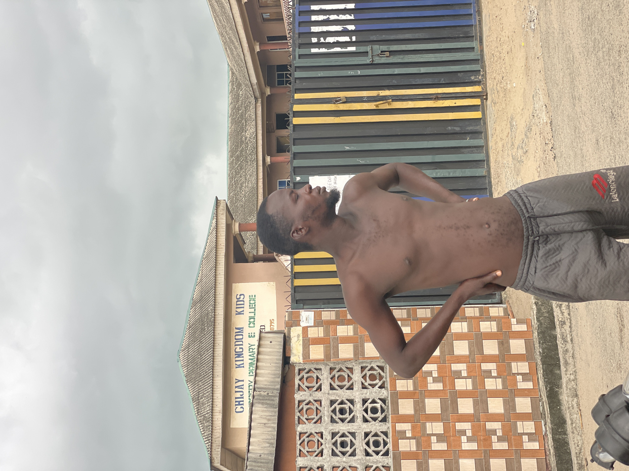 A shirtless man wearing gray shorts stands outside in Badagry, Nigeria, on an overcast day. He has dark skin and short black hair, with one hand on his hip and the other resting on his lower back. Behind him is a building with a sign that reads "CHIJAY KINGDOM KIDS primary & college". Next to the building is a decorated wall and a fence with colorful stripes. The ground is concrete.