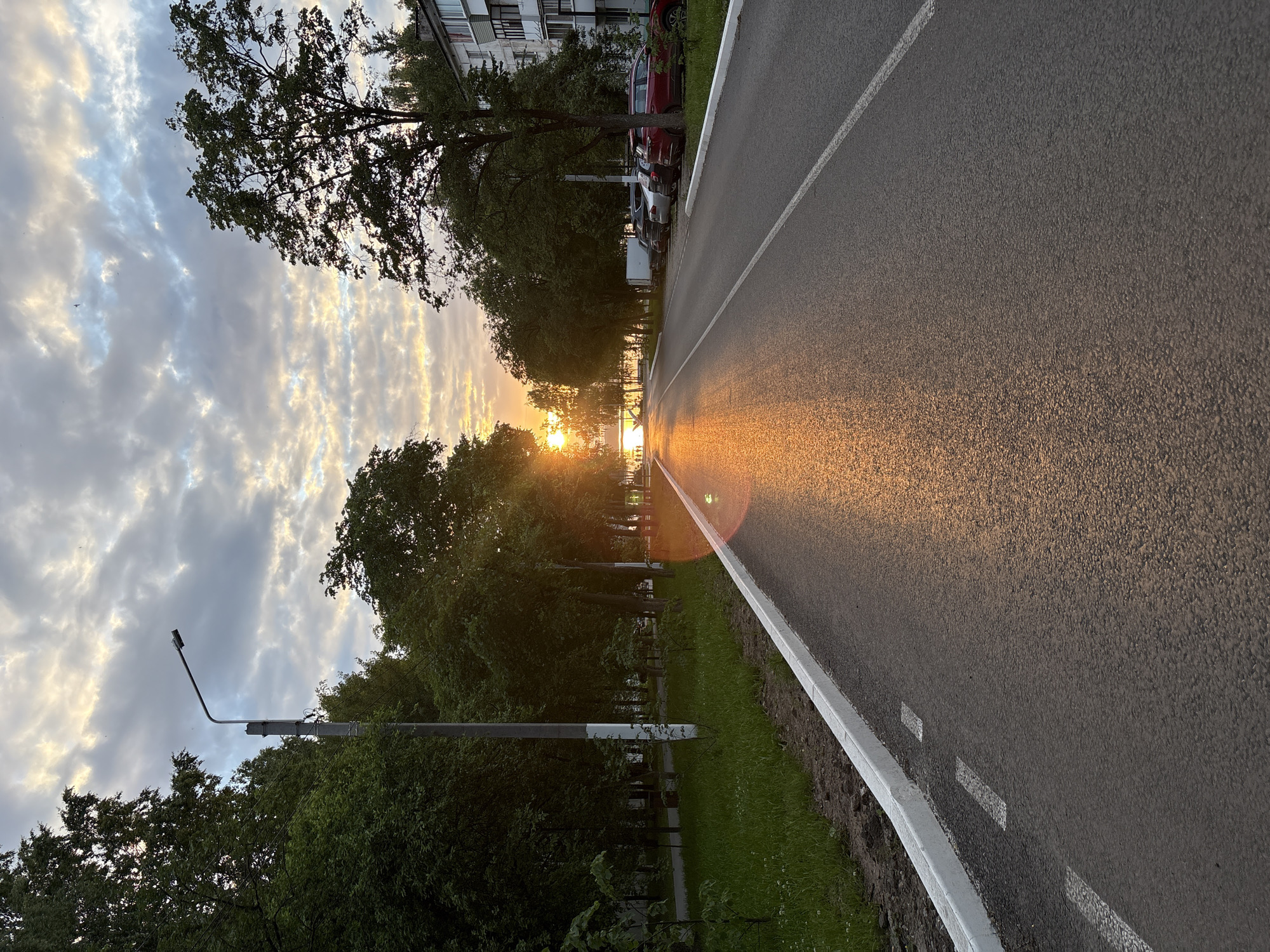 The image depicts a street scene in Chaikovsky, Russia, during sunset. The perspective is from a street facing the setting sun, which is visible in the distance, casting a warm, golden glow on the wet asphalt of the road. Trees line both sides of the street, with lush green foliage. On the right side of the road, parked cars are visible in front of a multi-story building. The sky is filled with dramatic, textured clouds, with hints of blue peeking through. A lamppost stands on the left side of the street. The overall atmosphere is serene and picturesque, capturing the beauty of a summer evening in Russia.
