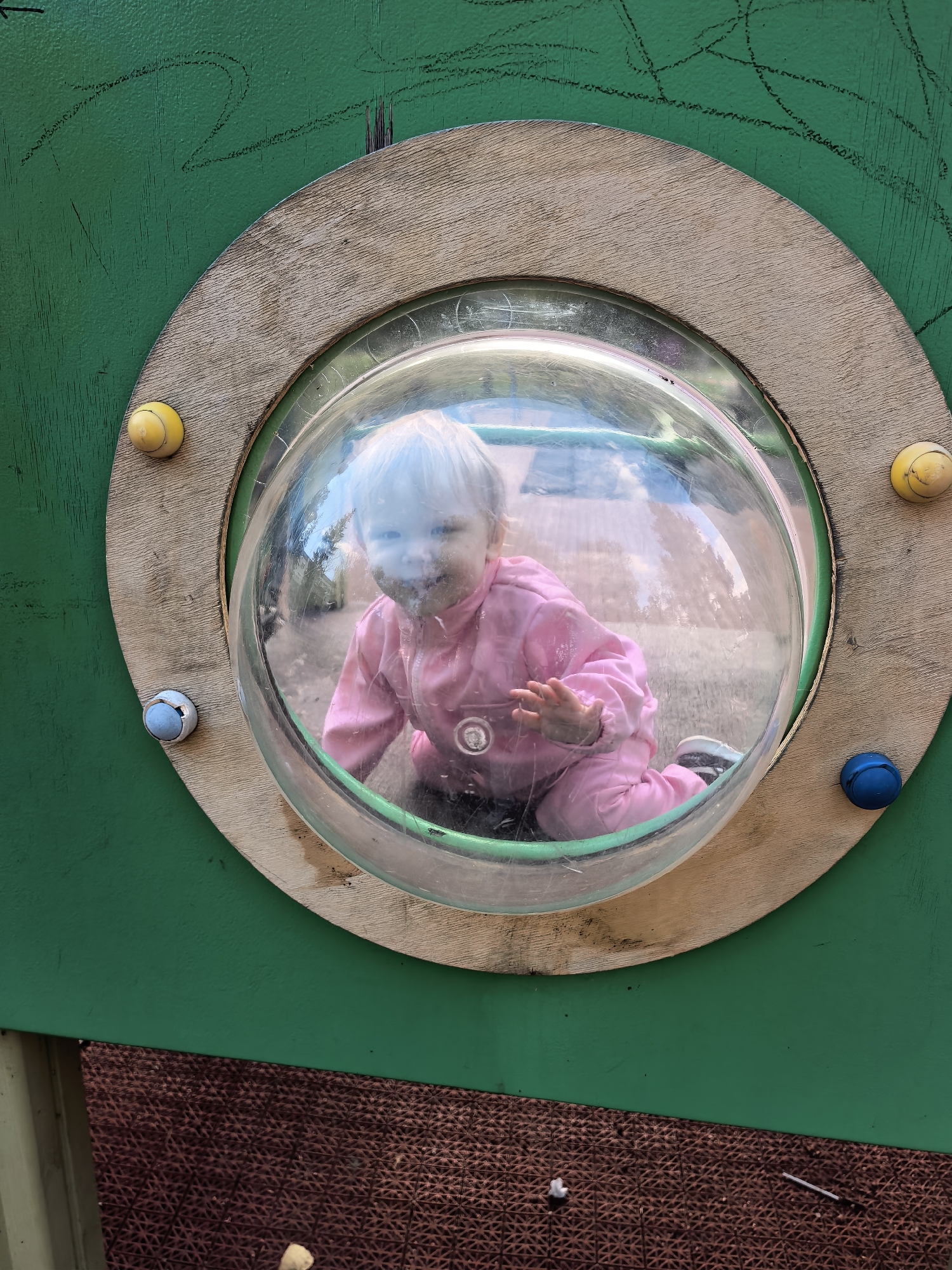 A child with blonde hair wearing a pink jumpsuit is crawling on a playground, viewed through a clear plastic dome. The dome is set into a green wooden structure. There are colorful balls attached to the structure around the dome: two yellow balls, one blue ball, and one light blue ball. The ground beneath the child appears to be dirt and gravel. The background outside the dome is blurred, showing hints of trees and sky. The overall scene suggests an outdoor play area. The location is identified as Sterlitamak, Russia.