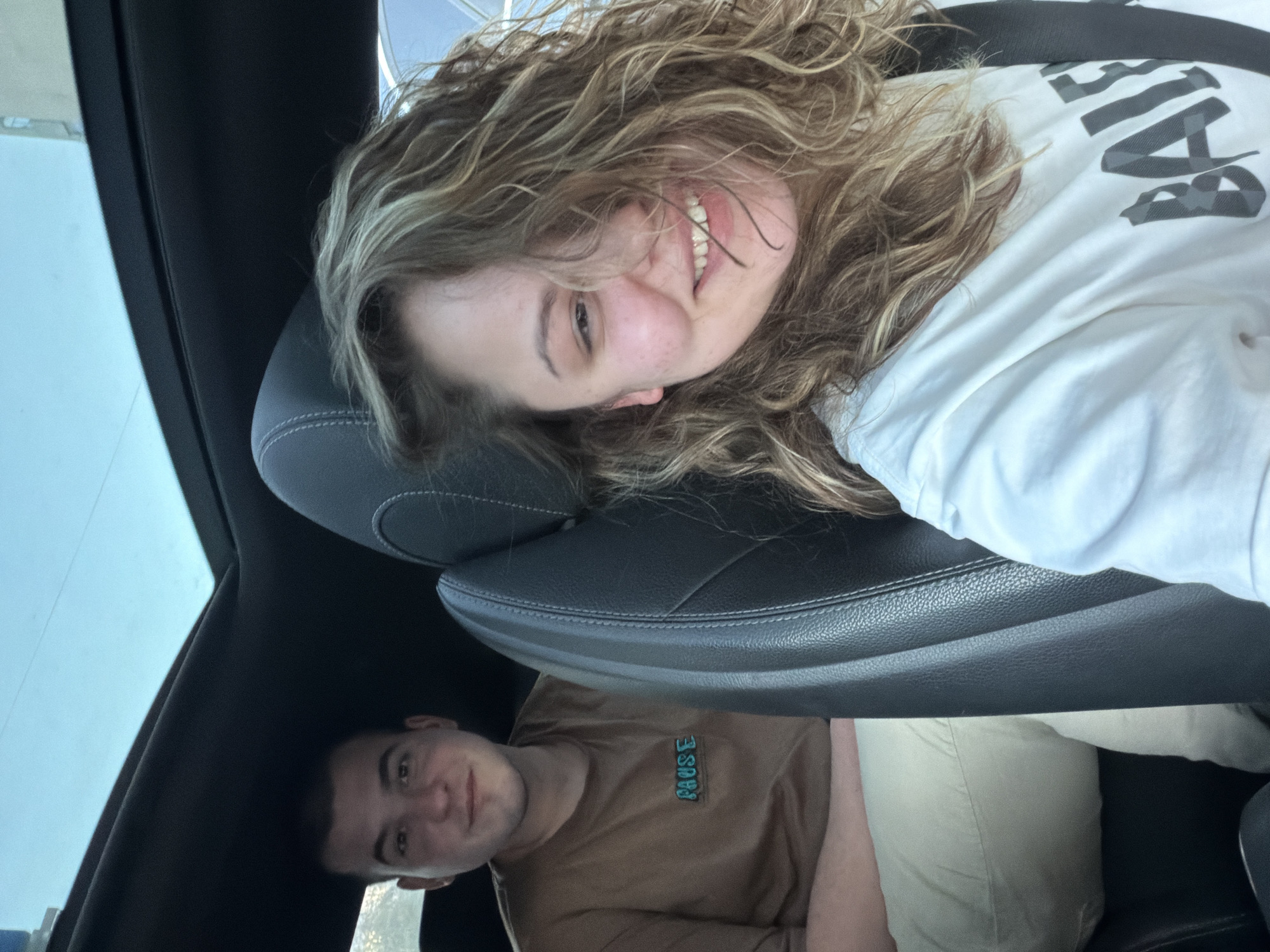 The image appears to be taken from inside a car. A female with curly, light brown hair is lying on a black leather car seat, smiling. She is wearing a white shirt with the word "Balenciaga" printed on it. Below her, another individual, a male, is visible. He has short dark hair and is wearing a brown t-shirt with the word "PAUSE" on it. He is also smiling. The photo is taken from an unusual angle, likely from the back seat looking forward. The car interior is dark, but outside, there is a glimpse of a light-colored building or structure. There are no specific details to place the location in Varna, Bulgaria, with certainty.