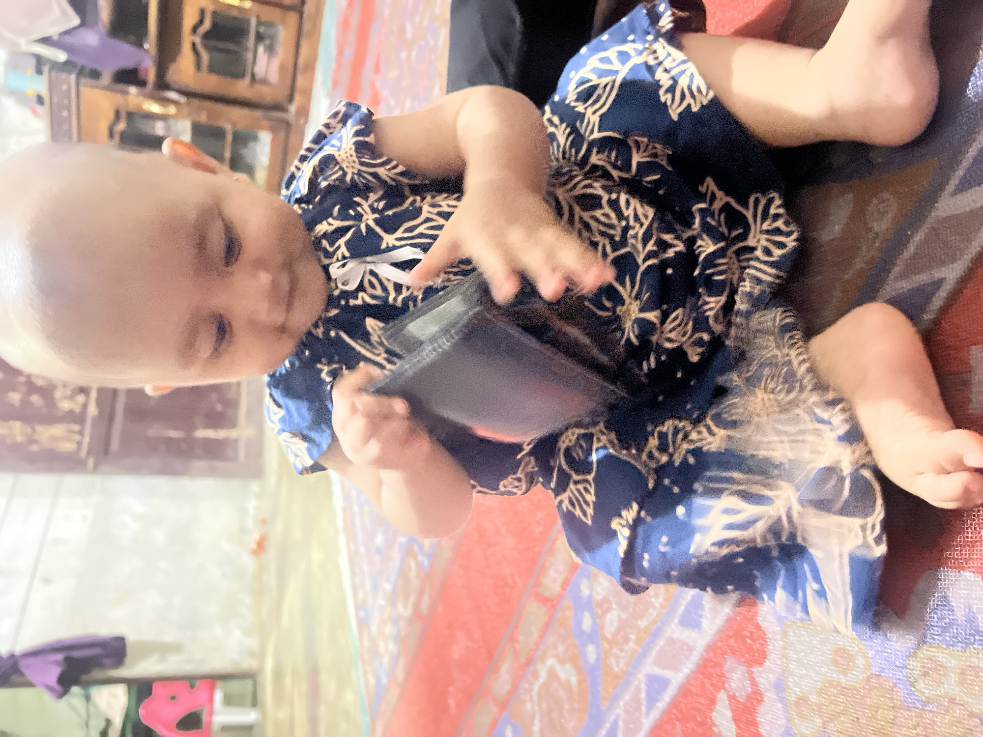 The image shows a baby girl wearing a dark blue dress with a white floral pattern and a white ribbon tied at the chest. She is sitting on a patterned rug with red, yellow, and blue accents, possibly in a home setting in Kilometer Delapan, Indonesia. The baby is holding a dark-colored object, possibly a wallet or a small book, and appears to be looking down at it. Her head is bald, and her eyes are open. The lighting suggests it is daytime. In the background, there are glimpses of wooden furniture, and on the right side of the image, there are parts of a rug or mat that is partially visible. The overall scene depicts a casual moment of a baby exploring an object.