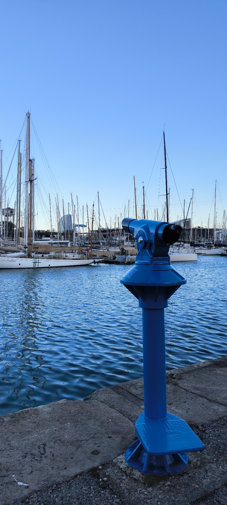 A standing blue coin-operated binocular telescope stands on a concrete block near the water, with a collection of docked sailboats and yachts in the background. The sky is a clear blue. The water in the foreground is a deep blue with small ripples. The telescope is positioned on a grey, rectangular concrete block. The sailboats and yachts have numerous masts reaching into the sky, with various rigging and sails furled or lowered. The background features a blurred cityscape.
