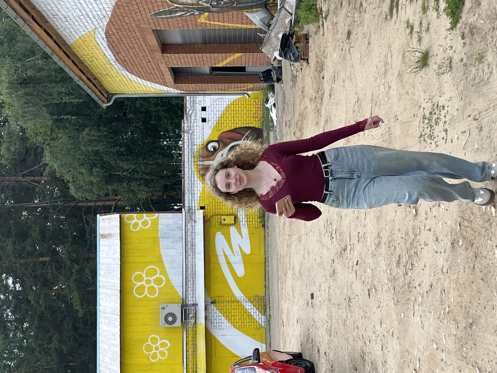 A young woman with curly blonde hair is walking towards the camera outdoors on a dirt path. She is wearing a dark red, long-sleeved shirt with lace trim at the neckline and light blue jeans. She is holding a smartphone in her right hand. Behind her, there are two buildings with colorful murals. The building on the left is yellow with white flower designs and an air conditioning unit. The building on the right is brick with a large mural featuring a dog's face. Trees are visible in the background. A red car is parked on the left side of the image. The scene appears to be in an industrial or semi-abandoned area.