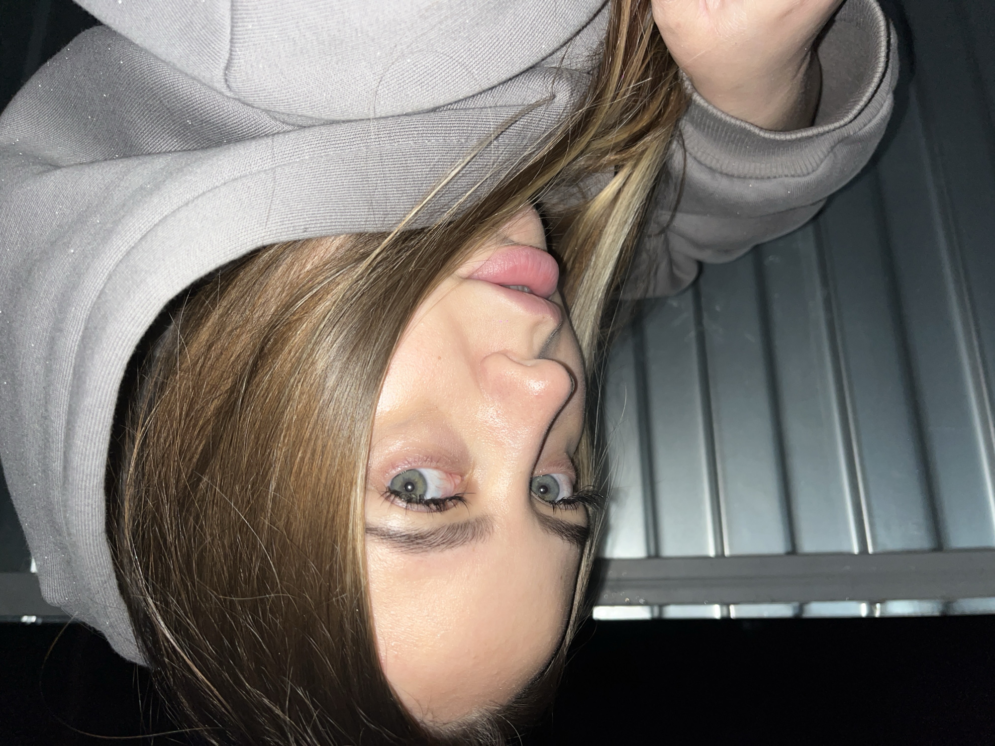 A person with fair skin, shoulder-length brown hair with blonde highlights, and light green eyes is photographed from an upside-down perspective. The person is wearing a light gray hooded sweatshirt. The background is a corrugated metal surface over a dark, unidentifiable area. The location is not discernable.