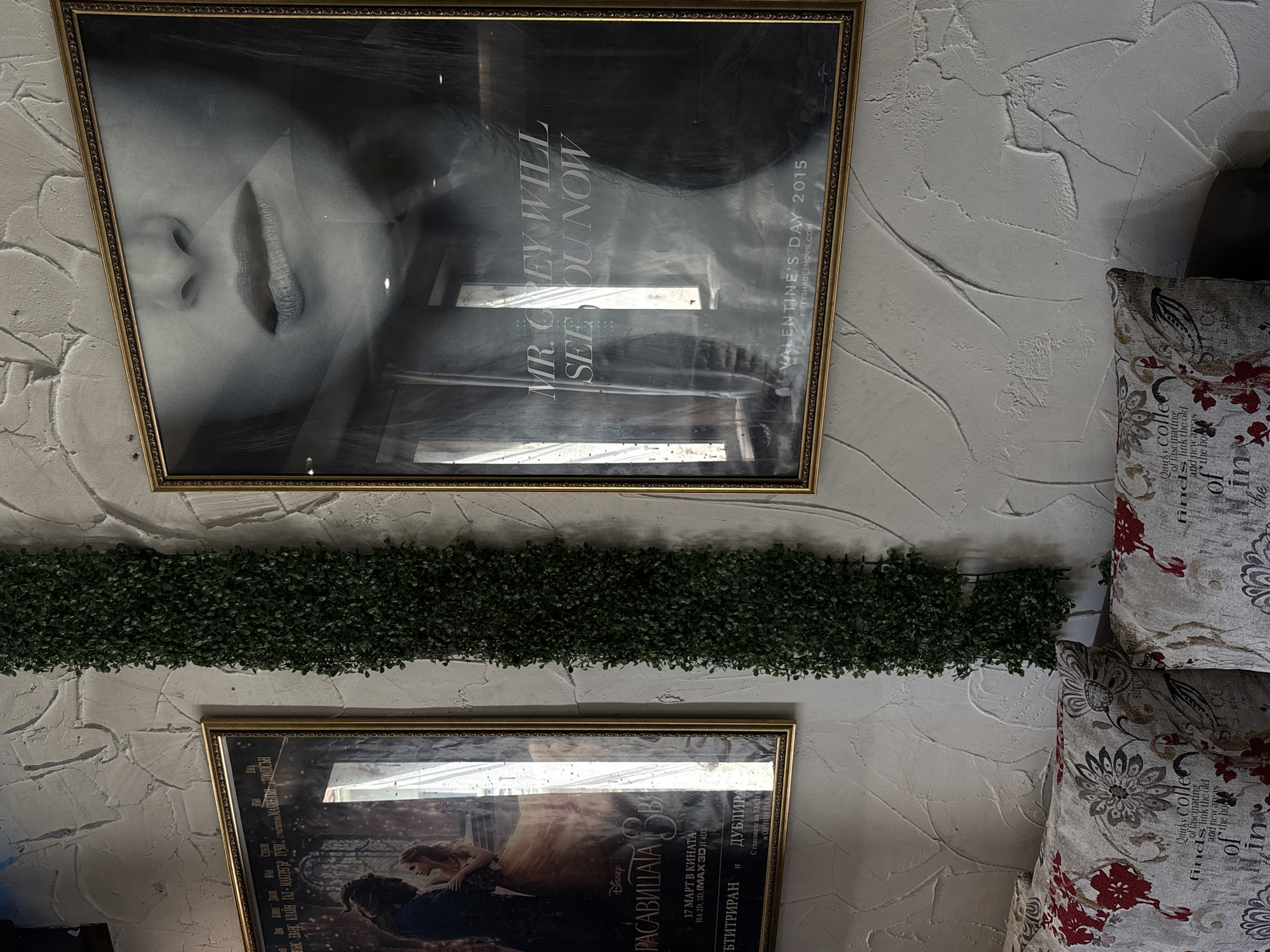 A close-up, slightly angled shot reveals a textured, off-white wall adorned with two framed movie posters and artificial greenery. Above, a poster for "Fifty Shades of Grey" features a partial face with the text "MR. GREY WILL SEE OU NOW" and "VALENTINE'S DAY 2015". Below it, separated by a strip of artificial greenery, is a poster for Disney's "Beauty and the Beast" (РАСАВИЦАТА ЗВЯ), in Cyrillic, with release date "17 МАРТ В КИНАТА". To the right, portions of two decorative throw pillows with floral and text patterns are visible. The location is Gotse Delchev, Bulgaria.