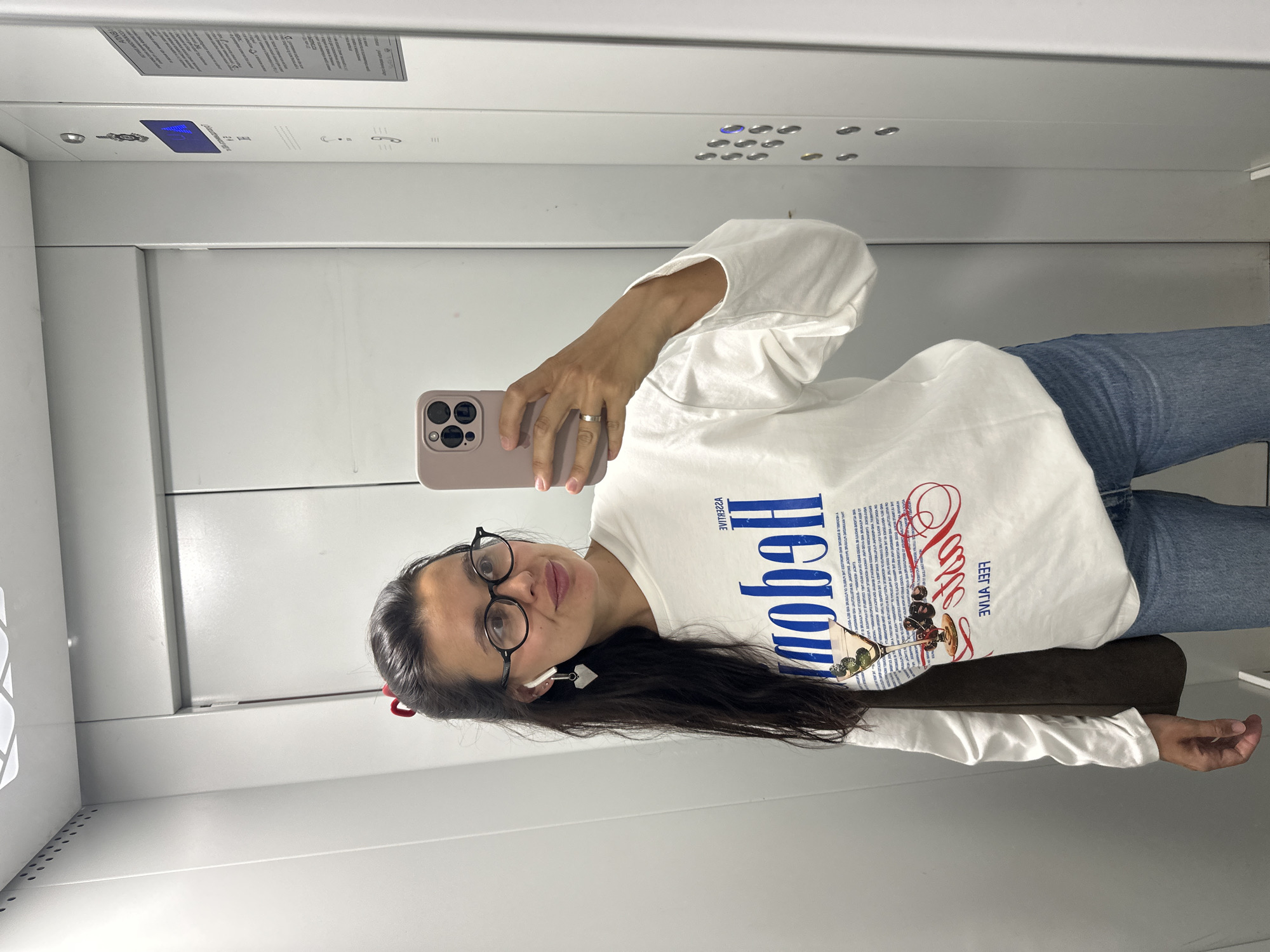 A woman is taking a mirror selfie in an elevator. She is wearing a white long-sleeved shirt with a graphic print and blue jeans. She has on round glasses and heart-shaped earrings. The elevator walls are silver and there are buttons on the right side of the frame. The image does not provide any information to suggest it is located in the city of Chaikovsky, Russia.