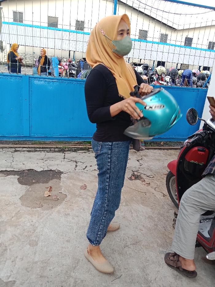 A woman is holding a blue motorcycle helmet in her right hand. She is wearing a beige headscarf and a black long-sleeved shirt. She is also wearing blue jeans and light brown flats. She is standing on a concrete surface. To the right of her is a red motorcycle and another person wearing khaki pants and brown sandals. The person appears to be mounting the motorcycle, but only the legs are visible. The background shows a blue metal gate and a row of parked motorcycles behind the gate. The scene is located in Tugu, Indonesia, at coordinates -6.973 latitude and 110.326 longitude. There are several other people standing in the background, who are not in focus. The environment is outdoors, with a cloudy sky in the background.