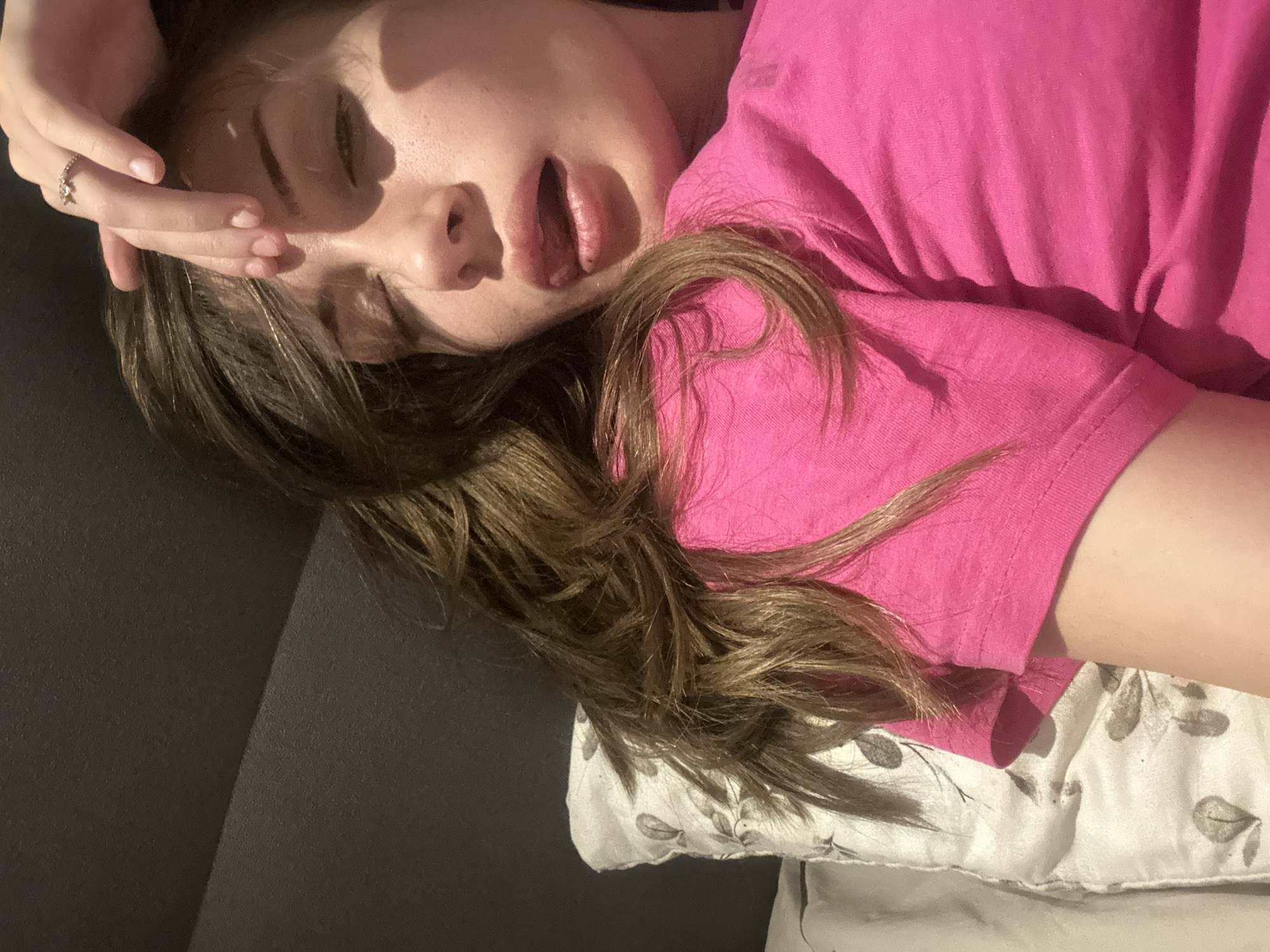 A young woman lies on her back on a gray sofa with a floral patterned pillow. She wears a pink t-shirt and has brown hair. Her eyes are closed and her mouth is open. Her right hand is raised to her forehead, and a ring is visible on one of her fingers. The scene is indoors, likely a private residence. There is no evidence to suggest the location is Burgas, Bulgaria.