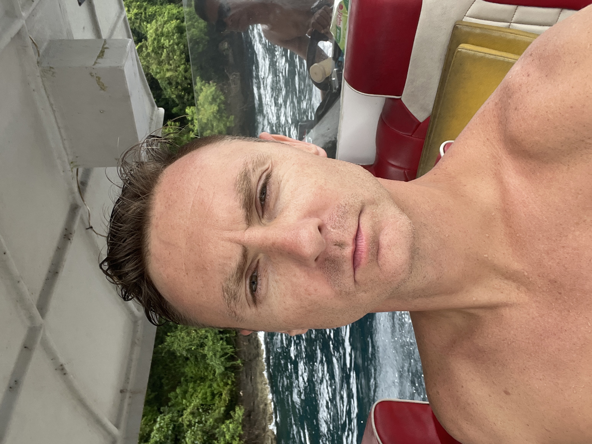 A close-up selfie of a shirtless man with wet brown hair, likely taken on a boat. Behind him is a body of water and green foliage. A red and white boat seat is partially visible. The location is likely in or near Kampung Toyapakeh, Indonesia, given the subject's proximity to the ocean and the type of vegetation visible. A reflection of another individual can be seen in a pane of glass.