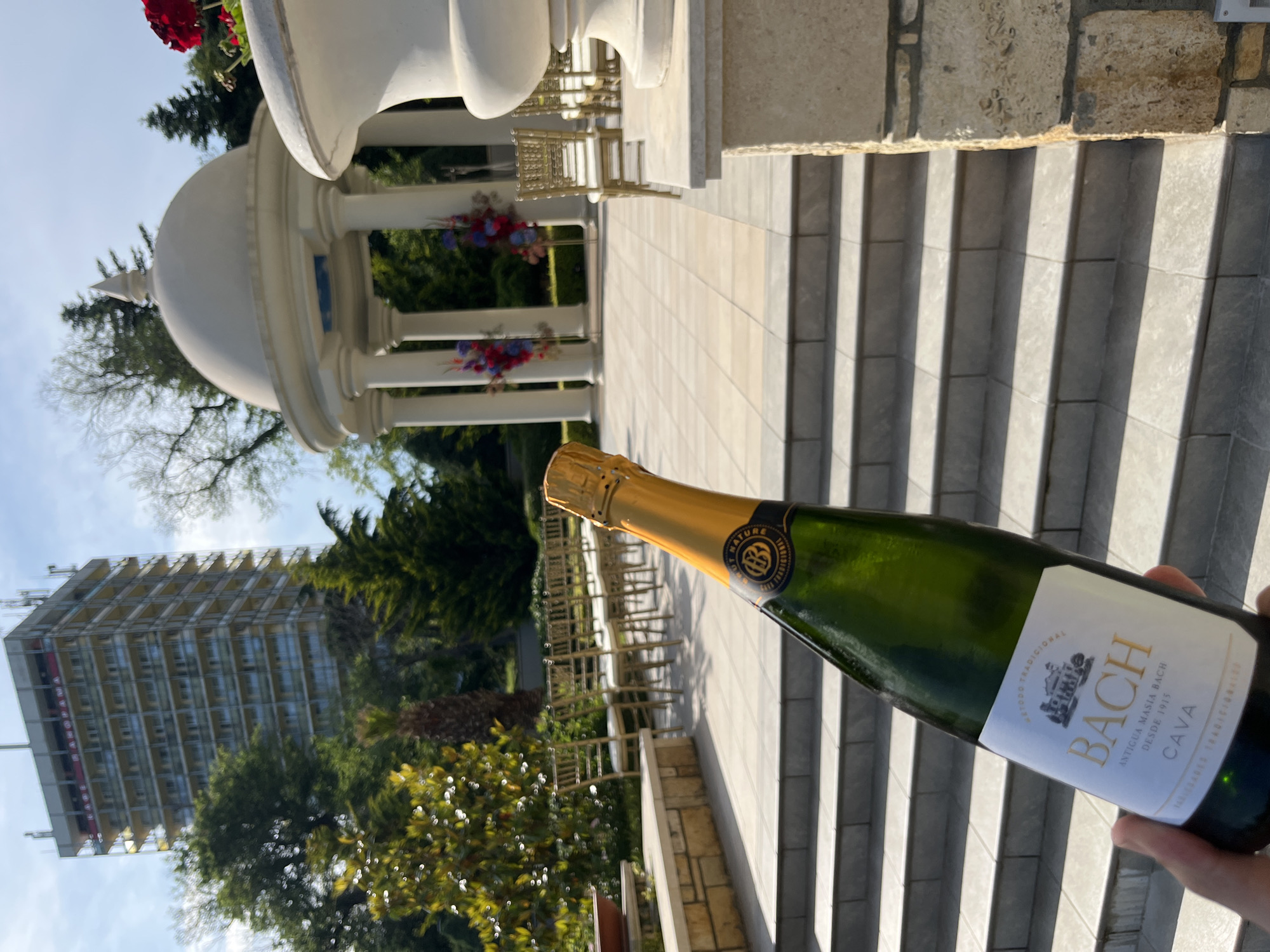 A hand holds a green glass bottle of "BACH CAVA" sparkling wine, featuring a white label with gold text reading "BACH," "ANTIGUA MASIA BACH DESDE 1915," and "CAVA." The bottle's neck is wrapped in gold foil with a dark blue label denoting "BRUT NATURE." The scene is an outdoor event space with stone-tiled steps leading down to a paved area. Golden Chiavari-style chairs with white cushions are arranged in rows, facing a white classical-style gazebo (rotunda) with a domed roof and several columns. Red and purple floral arrangements decorate the interior of the gazebo. Lush green trees and vegetation frame the midground. In the distant background, under a partly cloudy sky, a tall, multi-story building with numerous balconies is visible. A red sign near the top of this building clearly displays "КАФЕ ПАНОРАМА" (Cafe Panorama). This identifies the location as being in the city of Varna, Bulgaria, with the prominent building being the Panorama Hotel, and the event space likely part of its grounds or a nearby area such as the Sea Garden.