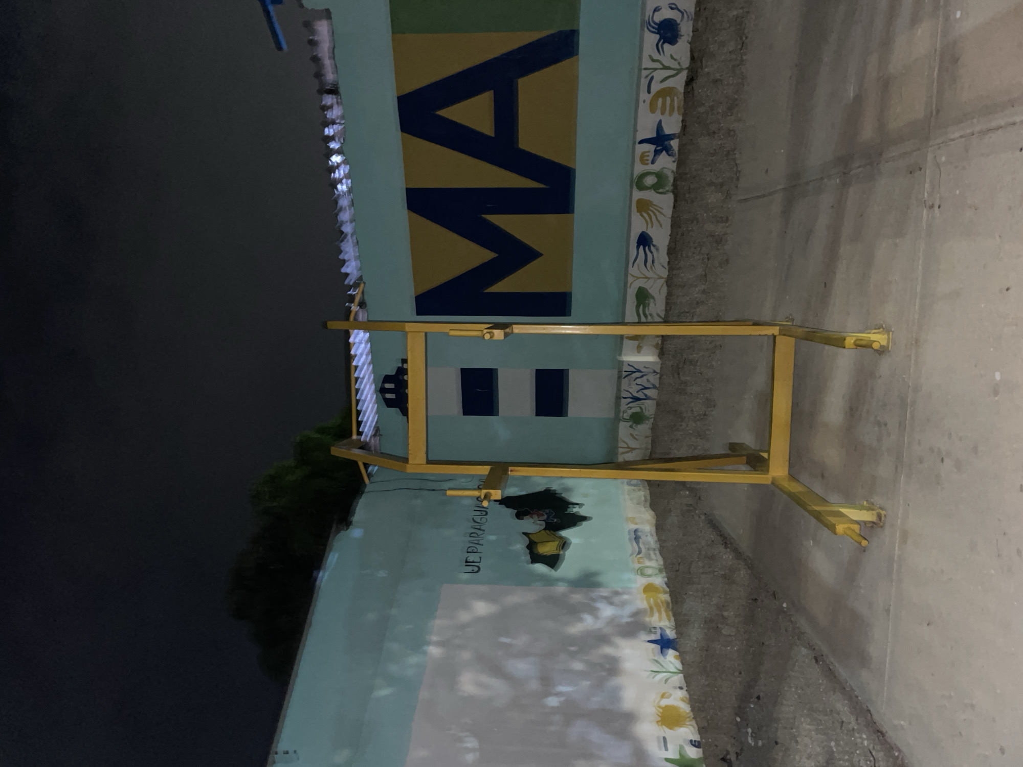 Captured at night, the image shows a building exterior in an urban setting, seemingly in Unknown, Unknown. The walls of the building are painted with marine-themed murals, featuring sea creatures like crabs and starfish. Bold, blue lettering is visible on one part of the building. A yellow metal frame or barrier stands near the building, possibly for construction or maintenance. The street or sidewalk next to the building is visible, showing signs of wear and use. The overall environment is urban, with darkness surrounding the immediate area.