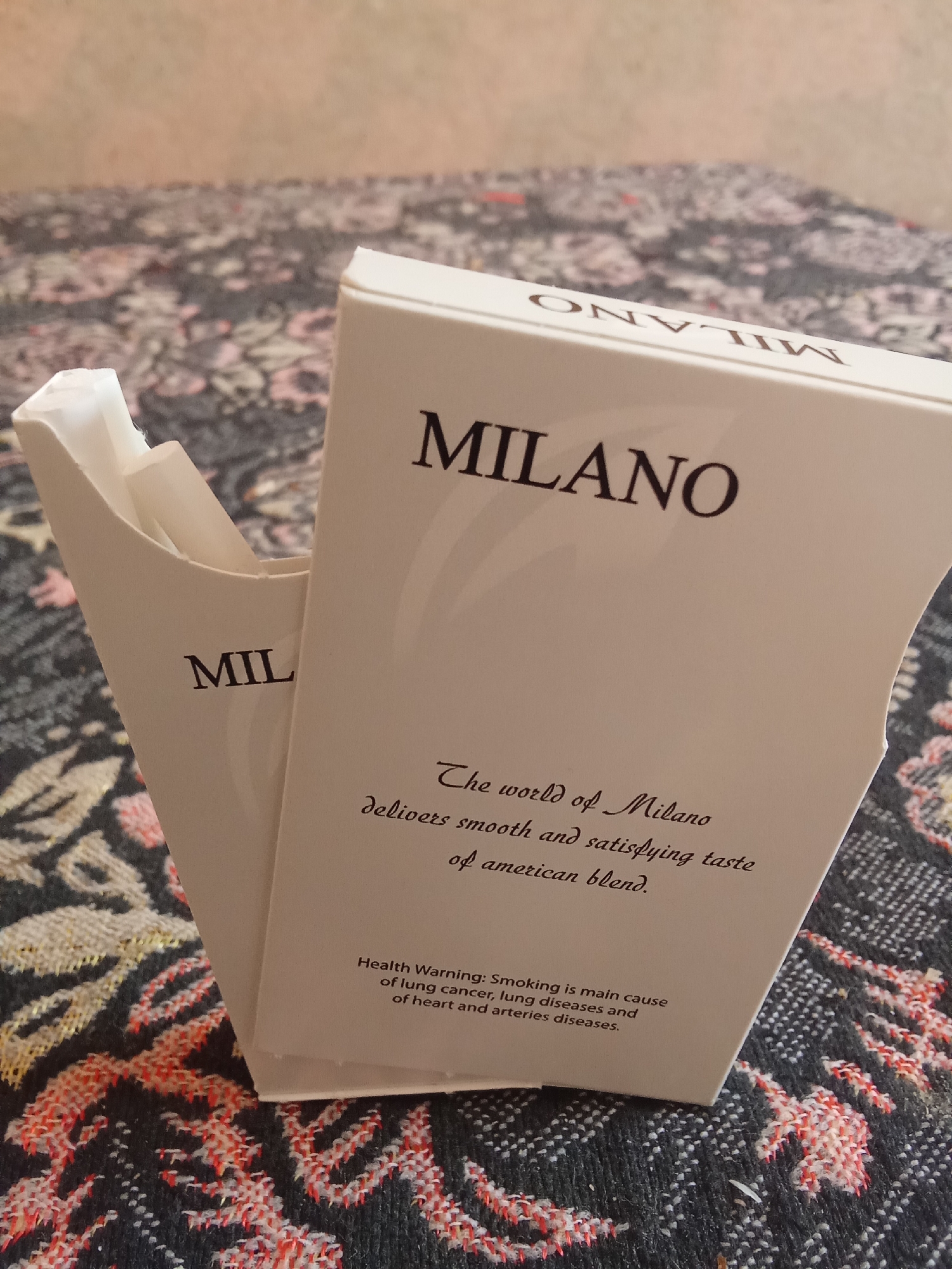 The image shows a partially opened pack of "MILANO" cigarettes on a patterned fabric surface. The pack is white with dark text and a curved graphic. The text on the pack reads "MILANO" prominently on the front. Below this, it says "The world of Milano delivers smooth and satisfying taste of american blend." There is also a health warning: "Health Warning: Smoking is main cause of lung cancer, lung diseases and of heart and arteries diseases." The cigarettes inside the pack are visible, some appearing to be filter tips. The fabric surface has a dark background with a dense pattern of pink, red, and cream floral or paisley-like designs. The background is blurred and appears to be a wall or similar surface. There are no people or actions depicted in the image. The image does not provide any information about its location in Fergana City, Uzbekistan.