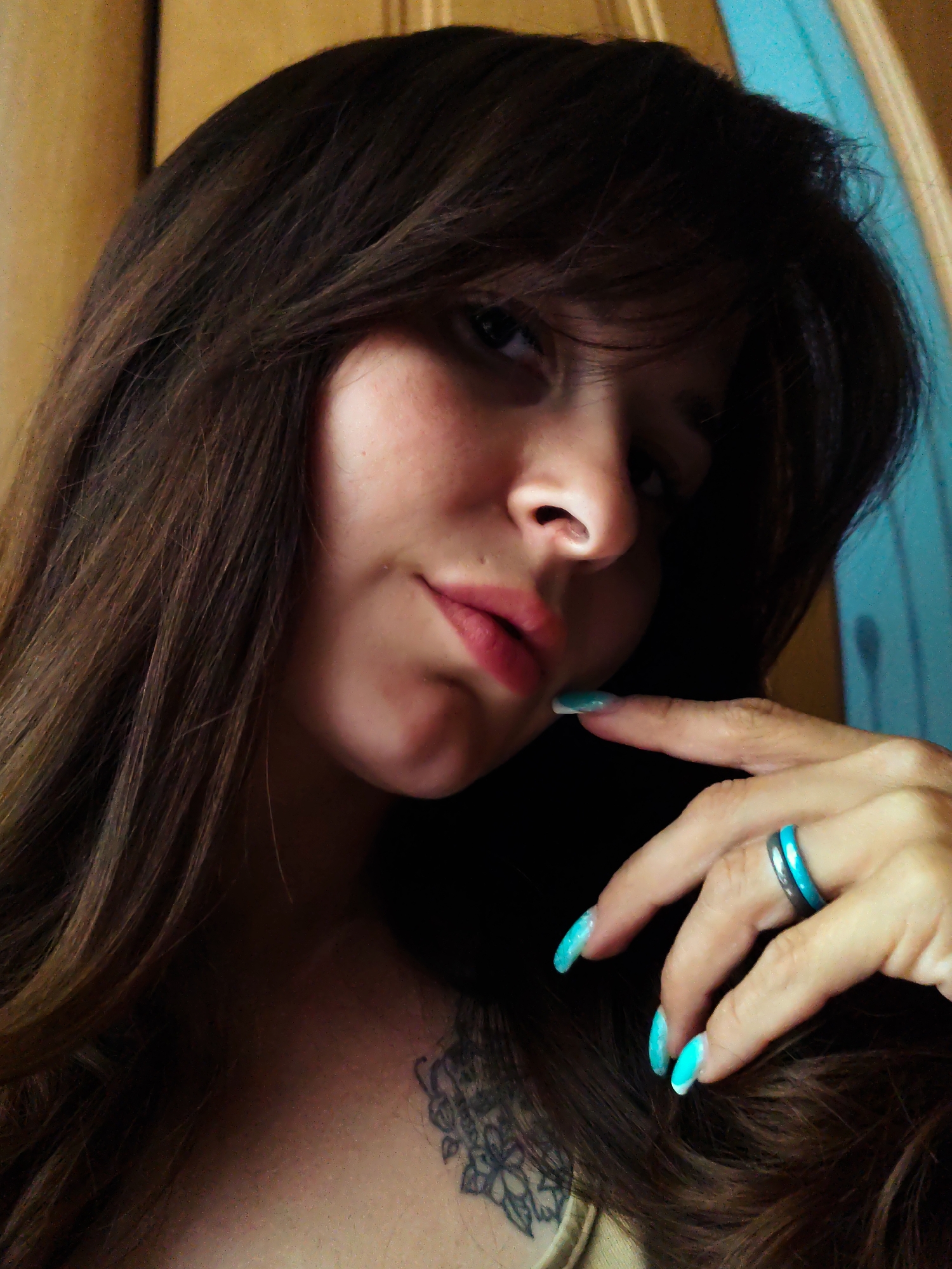 A close-up shot features a woman with long, wavy brown hair. She has fair skin, and her lips are tinted with a light pink color. She is wearing a ring with a turquoise band on her finger. Her nails are painted a matching turquoise color. A tattoo is visible on her chest. The background is blurred, with hints of brown and turquoise, which suggests an indoor environment. The city of Фоки, Russia, is not identifiable from the information contained within the image.