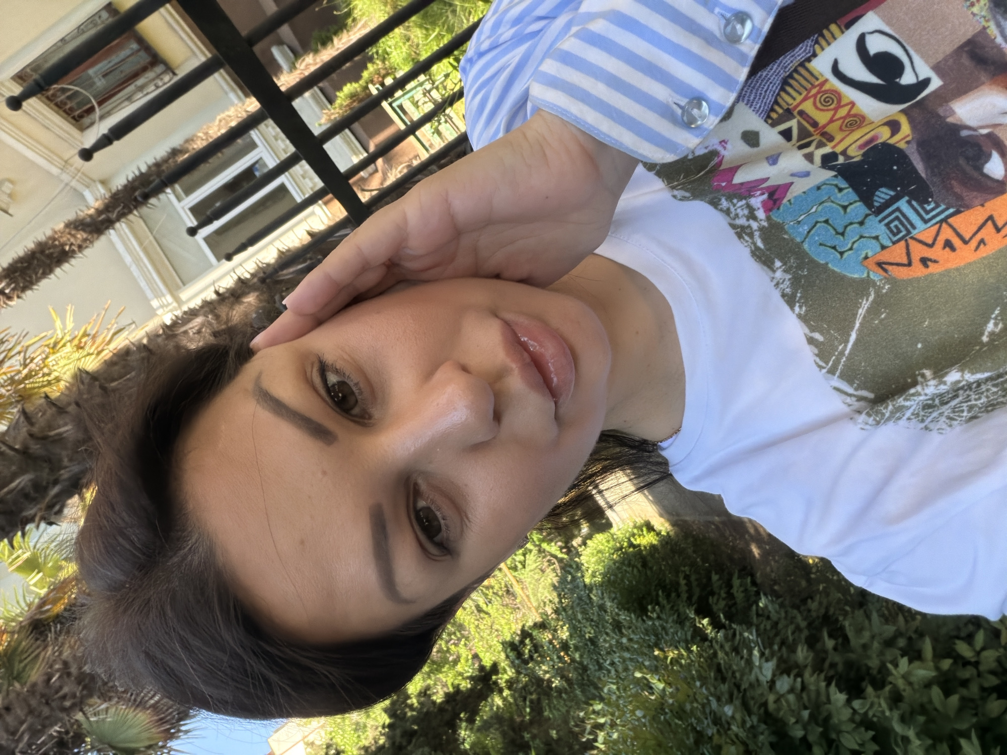 A female individual, appearing to be in her 30s or 40s, with dark brown hair and eyes, is depicted in a close-up outdoor self-portrait. She is wearing a white t-shirt with a multi-colored abstract collage print, partially covered by the sleeve of a blue and white striped long-sleeved shirt featuring clear buttons on the cuff. Her left hand rests gently on the side of her face, near her temple. The scene is illuminated by bright daylight. In the background, lush green subtropical vegetation, including visible palm fronds, is present. Behind this foliage, a light-colored building with white window frames and dark security bars is partially visible. A black metal fence with spherical finials extends across the midground. The presence of subtropical flora, particularly palm trees, is consistent with the climate and typical environment found in Sochi, Russia.