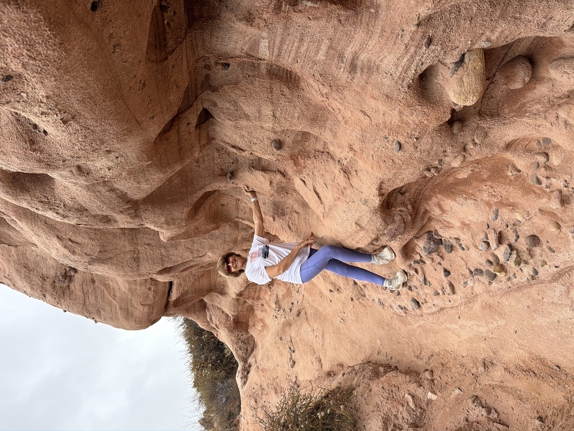 A woman is captured mid-climb on a sandstone rock formation. She is wearing a white t-shirt with a graphic on the front, purple leggings, and athletic shoes. The woman has on sunglasses and is smiling. Her right arm is extended upward, gripping the rock, while her left hand is on her hip. The rock formation has a rough, textured surface with visible layers and embedded stones. The scene appears to be outdoors, with a cloudy sky visible in the background. The ground around the rock formation is sandy and littered with small pebbles. There is some dry brush or vegetation growing at the base of the formation. The overall impression is one of an active and enjoyable outdoor pursuit. The image is oriented sideways. No visible text other than what appears to be a graphic on the woman's shirt.