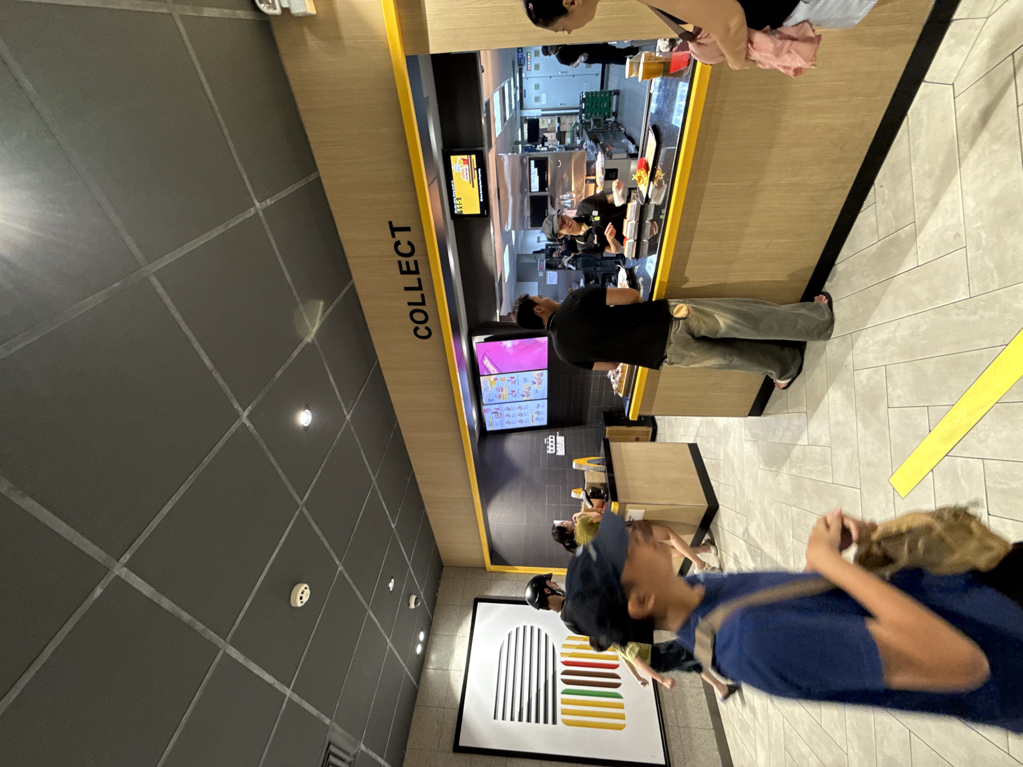 The image depicts the interior of a fast-food restaurant, likely a McDonald's due to the branding and menu boards, in Nha Trang, Vietnam. A group of people are queuing to collect their orders. The restaurant features a modern design with a grey tiled ceiling, wooden paneling, and yellow accents. A large abstract artwork is visible on the left wall. The main action focuses on the "COLLECT" counter where a staff member is handing food to a customer. The overall atmosphere is busy, typical of a lunchtime rush.