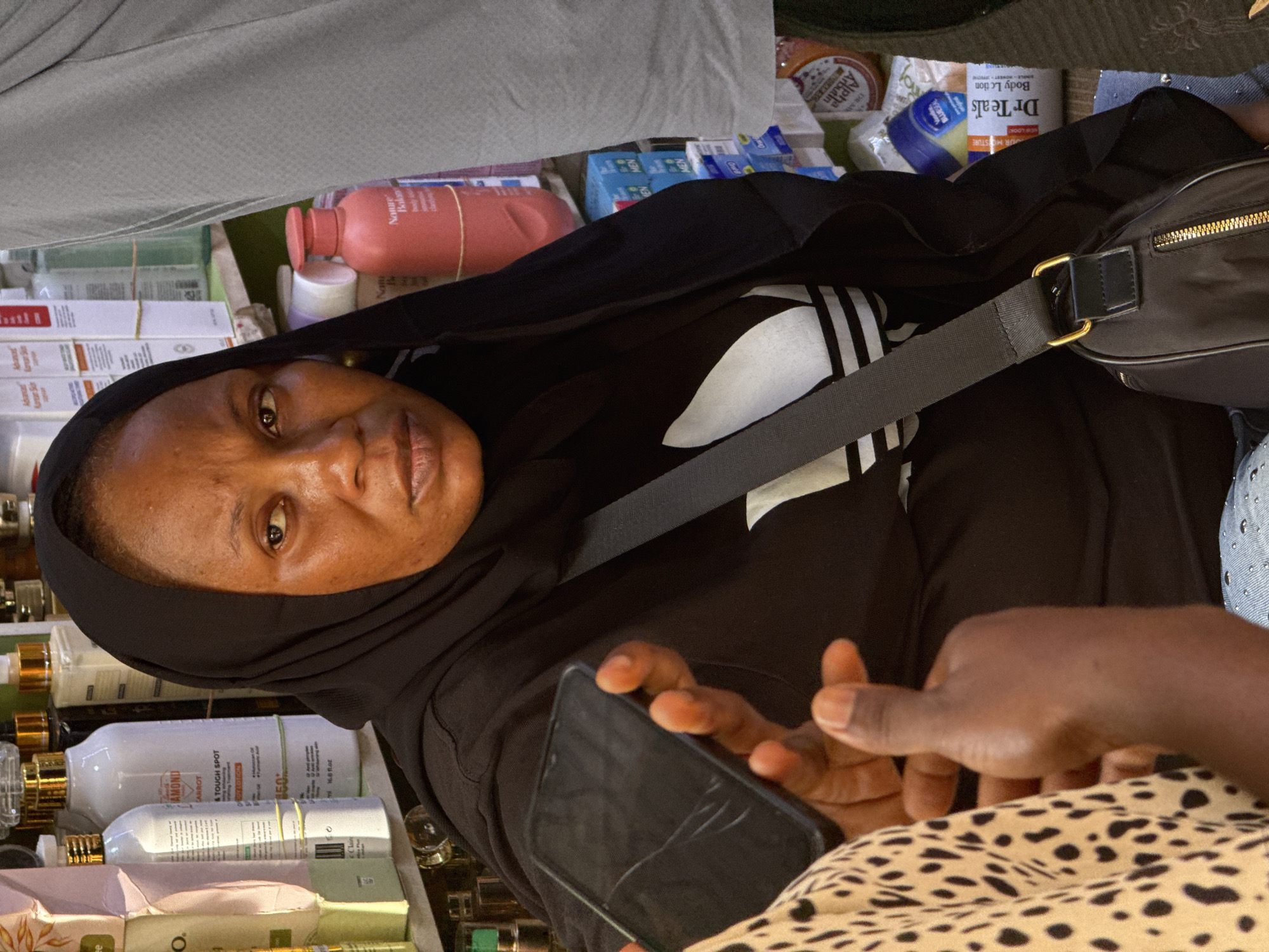 A woman wearing a black hijab and an Adidas-branded top is pictured indoors, possibly in a retail environment. She is holding a smartphone with a cracked screen. In the background, there are various beauty and skincare products, including lotions and creams. Some visible brands include "Dr Teal's," "Nature's Body" and "Claire Diamond Carrot". Another person wearing an animal print top and holding out a hand is partially visible.