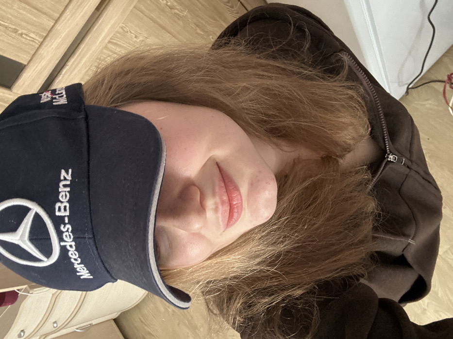 The image depicts a female individual, photographed from a high-angle, overhead perspective, in what appears to be an indoor setting in Nizhnekamsk, Russia. The individual possesses light brown hair and is wearing a dark blue baseball cap. The cap features the Mercedes-Benz logo and text embroidered in white on the front, with additional white text, partially visible as "West McLare," on its right side. The brim of the cap obscures the subject's eyes. Her lips are slightly parted, suggesting a subtle smile, and a minor blemish is visible on her chin. She is dressed in a dark brown zippered hooded garment, with the zipper partially unfastened. The background includes a light-colored, vertically ribbed fabric surface on the left, a light wood-toned door or wall panel on the right, and a white object, possibly an appliance or furniture piece, with dark cables extending from it visible on a light-colored floor in the lower right portion of the frame.