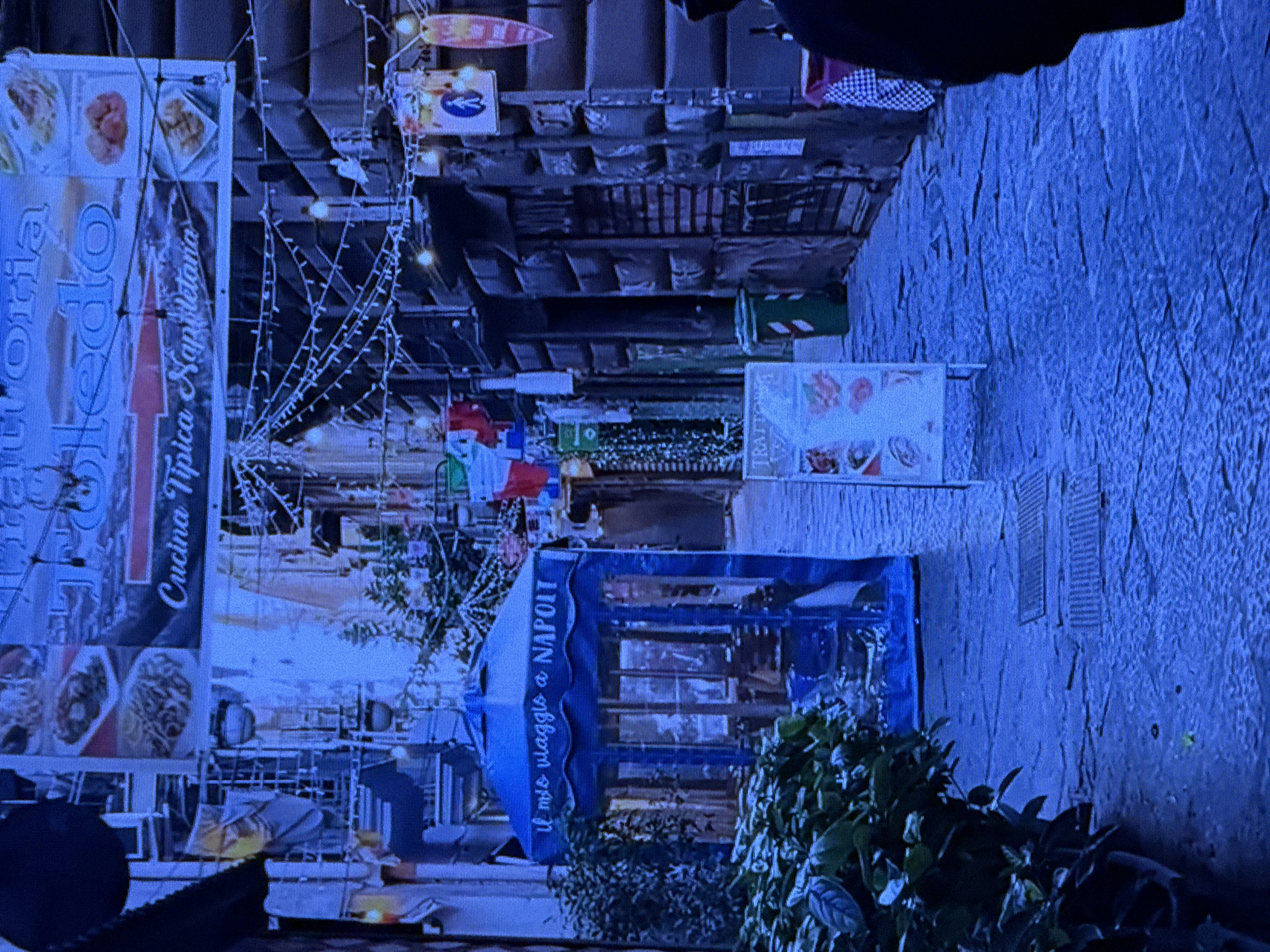 This media file depicts a nighttime scene on a cobblestone street, likely in a Mediterranean or European location, given the visible text. The lighting suggests it is evening or night, with an atmosphere illuminated by strings of festive lights.
The scene is dominated by various objects and structures. On the left, a large banner displays images of food and text in Italian, including "Trattoria," "Napolitana," and "Cucina Tipica." Adjacent to this, a blue awning with text in Italian, "Il mio viaggio a Napoli," is visible. Further down the street, other stalls or shops are present, some with colorful awnings and signs. In the background, buildings with multiple stories and balconies are discernible, adorned with more lights and decorations. A green trash bin with red and white stripes is also present.
The overall impression is that of a lively street market or a street lined with restaurants and shops, possibly during a festive season. The presence of outdoor seating and the festive lighting contribute to a welcoming and vibrant ambiance. There are no discernible people in the frame, but the setup implies activity.
The image is characterized by a prominent blue hue, likely due to the lighting conditions and the dominant colors used in the scene. The cobblestone texture of the street is also a notable detail, suggesting an older, perhaps historic, urban setting. While the media file is presented with a Dubai, United Arab Emirates context, the visible text and architectural style do not strongly support this location and are more indicative of a European setting, specifically Italy.