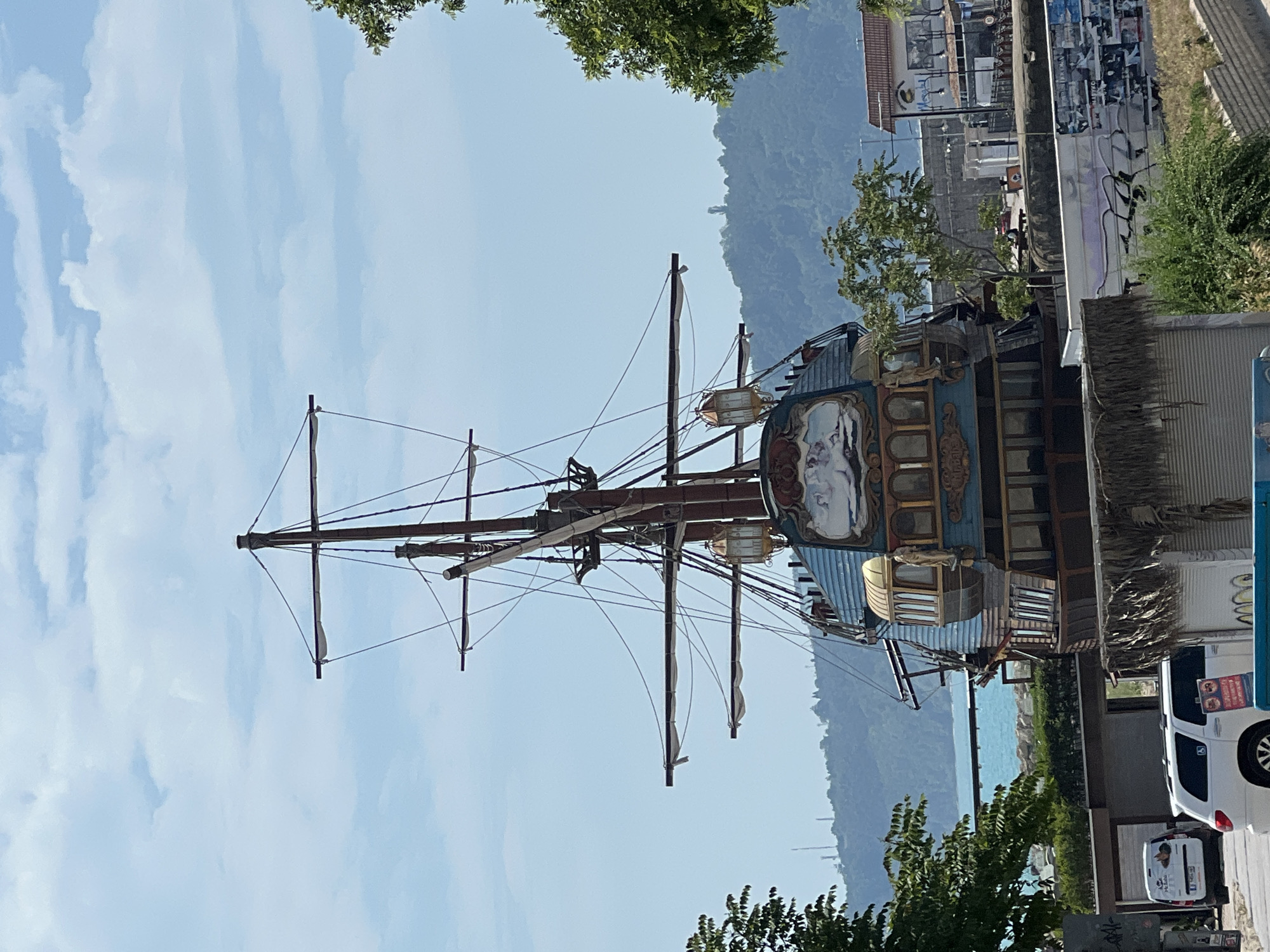 The image captures a unique, decorative ship-like structure, likely a themed restaurant or attraction, situated on land. This prominent subject features multiple tall masts with extensive rigging, and its ornate hull includes a large painted mural depicting a figure on its side, along with several detailed cabin-like windows and lamps.
In the background, a large, tree-covered mountain range rises under a bright, clear blue sky dotted with soft white clouds. A calm, turquoise sea is visible in the distant background, beyond the mountain range.
To the right of the ship structure, the scene includes urban elements: various buildings, one of which has a wall covered in posters or graffiti. Two white vehicles, a larger SUV/minivan and a smaller car, are partially visible on the right side of the frame. Green trees and other foliage are scattered throughout, contributing to a vibrant, natural feel amidst the man-made structures.
The photograph was taken during daytime, indicated by the bright sunlight and clear weather. No specific human activity is discernibly taking place. While some text may be present on the vehicles and buildings, it is too small or obscured to be legible. The overall setting strongly suggests a coastal city or resort area.