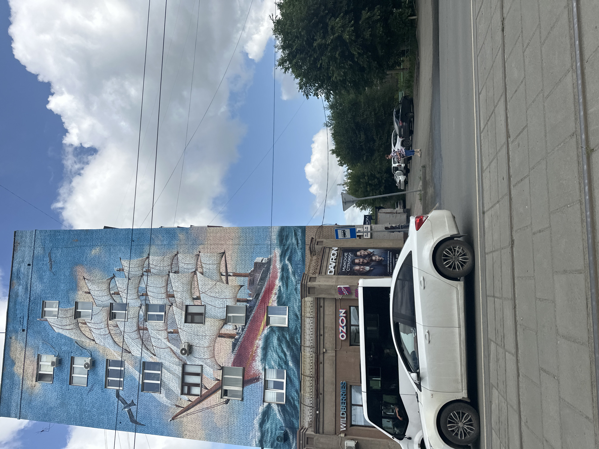 The image shows a multi-story brick building with a large mural of a sailing ship on its facade, located in Orsk, Russia. The mural depicts a red-hulled ship with white sails against a blue, wave-filled ocean and sky. Below the mural, on the ground floor, are storefronts including "WILDBERRIES" and "OZON," as well as an advertisement for "DAVRON" men's haircuts. A white passenger van and a white sedan are parked on the street in front of the building. A person in a patterned shirt and blue jeans is standing in a parking area to the right of the building, near several other parked cars and a densely wooded area. Power lines are visible against the sky. The street in the foreground has tram tracks.