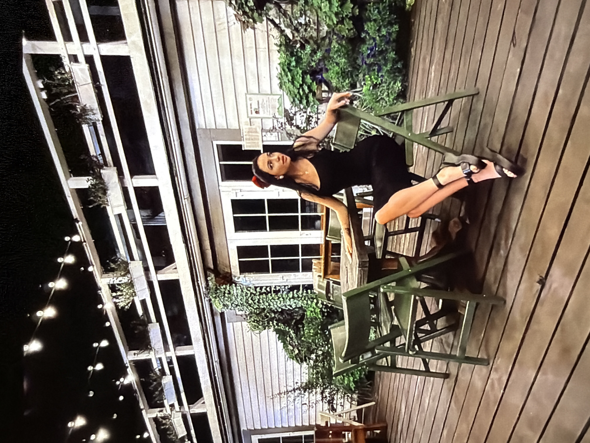 A woman with long dark hair, wearing a black spaghetti strap dress and black platform heels, sits on a green folding chair at a wooden table on a wooden deck. She is wearing a red bow in her hair and white earrings. The deck is surrounded by wooden railings and planters with greenery. In the background, there is a building with white horizontal siding and several windows. String lights are visible overhead. The scene appears to be an outdoor seating area at night.