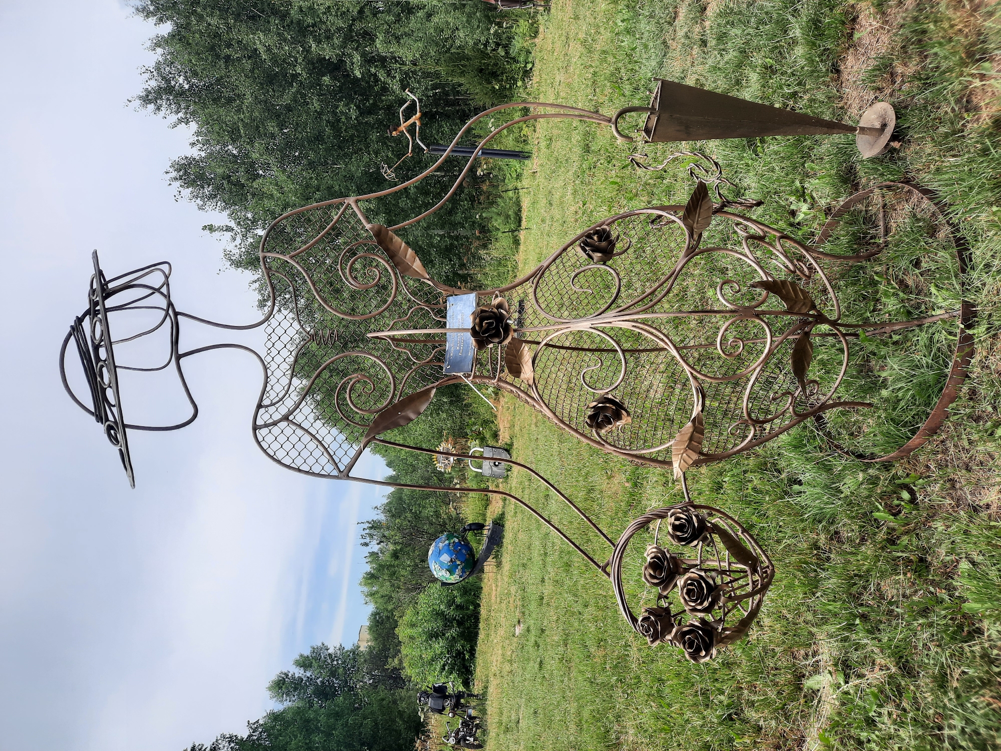 A life-sized metal sculpture of a woman stands in a grassy, outdoor area. The sculpture is made of wire and mesh, with decorative metal leaves and roses adorning its body. The woman is wearing a wide-brimmed hat and holding a basket of roses in her left arm and a cone-shaped object, possibly an umbrella stand, in her right hand. In the background, there are trees, a globe on a stand, a motorcycle with a robot figure, and a large padlock sculpture. The sky is overcast. The sculpture is located in an open field that appears to be part of a park or sculpture garden.