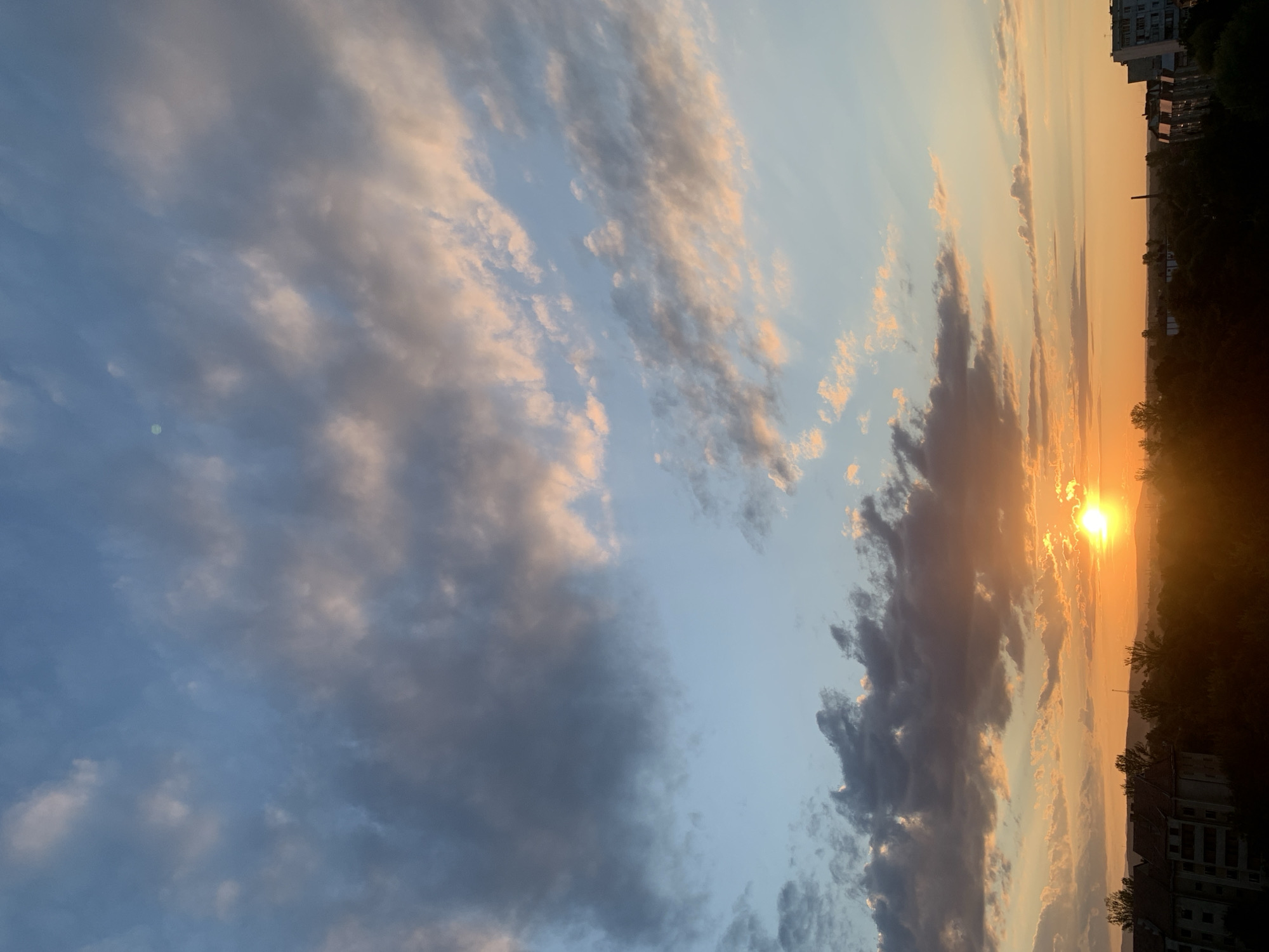 The image captures a sunset over the city of Kolomyia, Ukraine. The sky is filled with a mixture of blue and orange hues, with clouds of varying shades of grey and pink. The sun, a bright orange orb, is setting near the horizon, casting a warm glow over the landscape. In the foreground, the silhouettes of buildings and trees are visible, suggesting an urban or semi-urban environment. The architecture of the buildings appears to be typical of Eastern European cities. The overall impression is one of a serene and picturesque evening scene.
