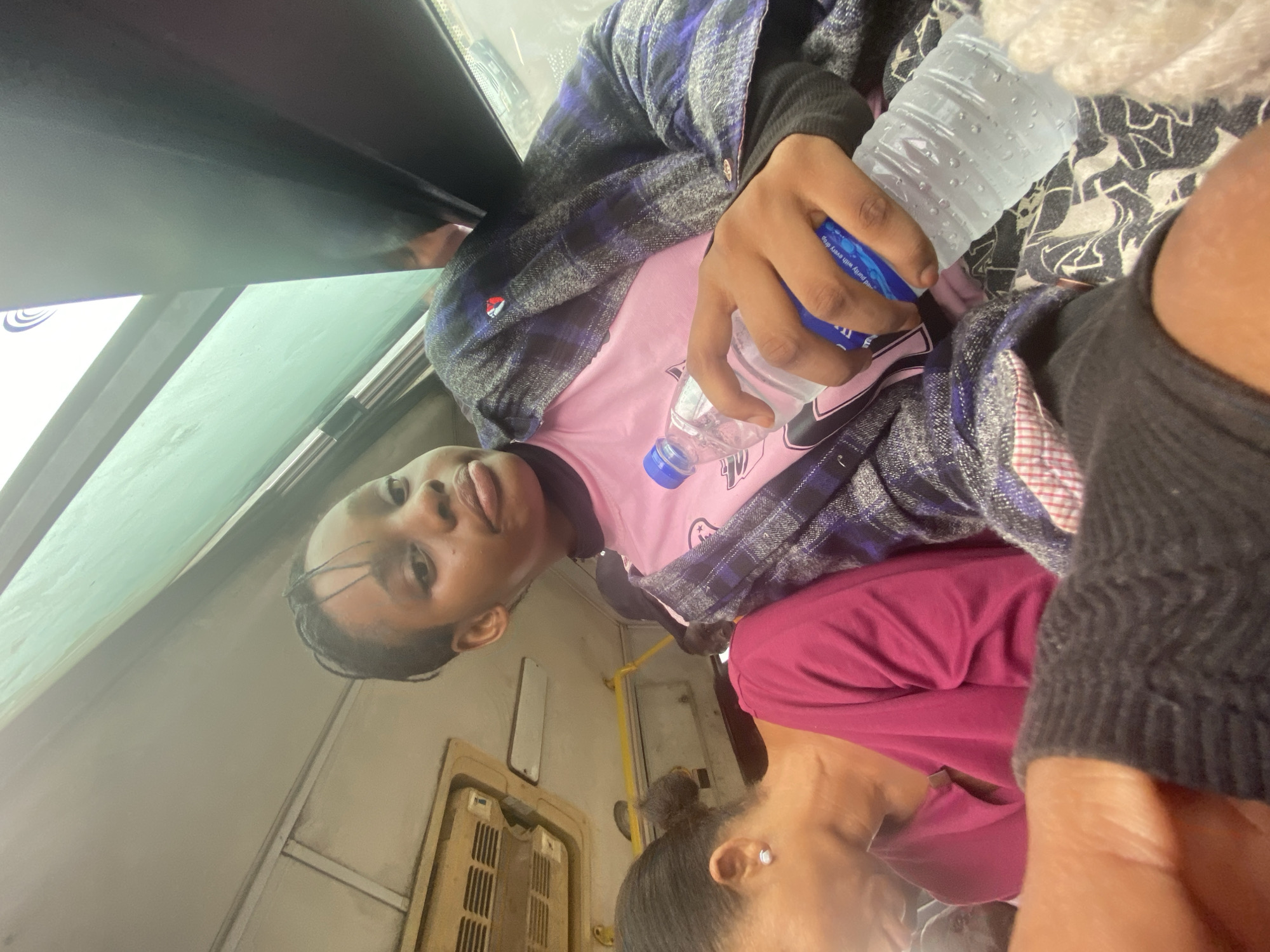 A medium shot taken inside a train car, location Unknown, Unknown, captures a young Black person in a pink t-shirt and a plaid button-down shirt, holding a water bottle. The scene includes additional unidentified individuals partially visible on either side of the primary subject. A window is to the left, and a door with ventilation slots is in the background.