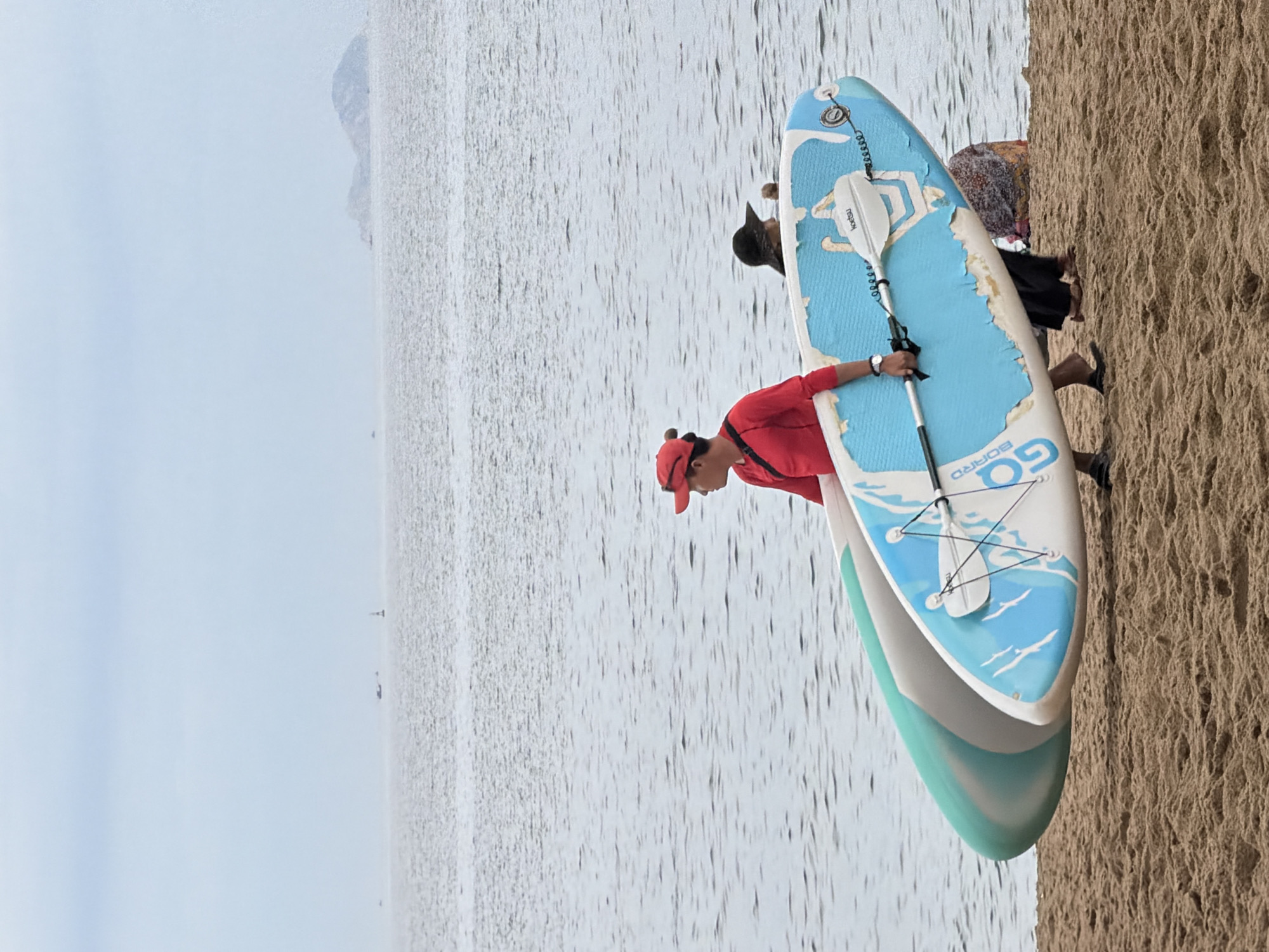 In Nha Trang, Vietnam, two stand-up paddleboards rest on a sandy beach, positioned at an angle. The foreground features the textured surface of the sand, leading towards the water. The ocean stretches out beyond, appearing slightly choppy under an overcast sky. In the distance, a hazy landmass with a rounded top is visible on the horizon, with a few small boats scattered across the water.
Two individuals are depicted in the middle ground, near the water's edge. A person wearing a red cap and a red long-sleeved shirt is carrying one of the paddleboards. This individual is facing right and appears to be walking. Another person, wearing a dark hat and a patterned garment, is partially obscured behind the paddleboards and appears to be standing or walking in the water or near it. The paddleboards themselves are distinct: one is predominantly turquoise, and the other is white with a large blue design and white lettering that reads "GO BOARD." A paddle is secured to the blue and white board. The overall impression is a casual scene by the sea, possibly a recreational activity about to begin or conclude.